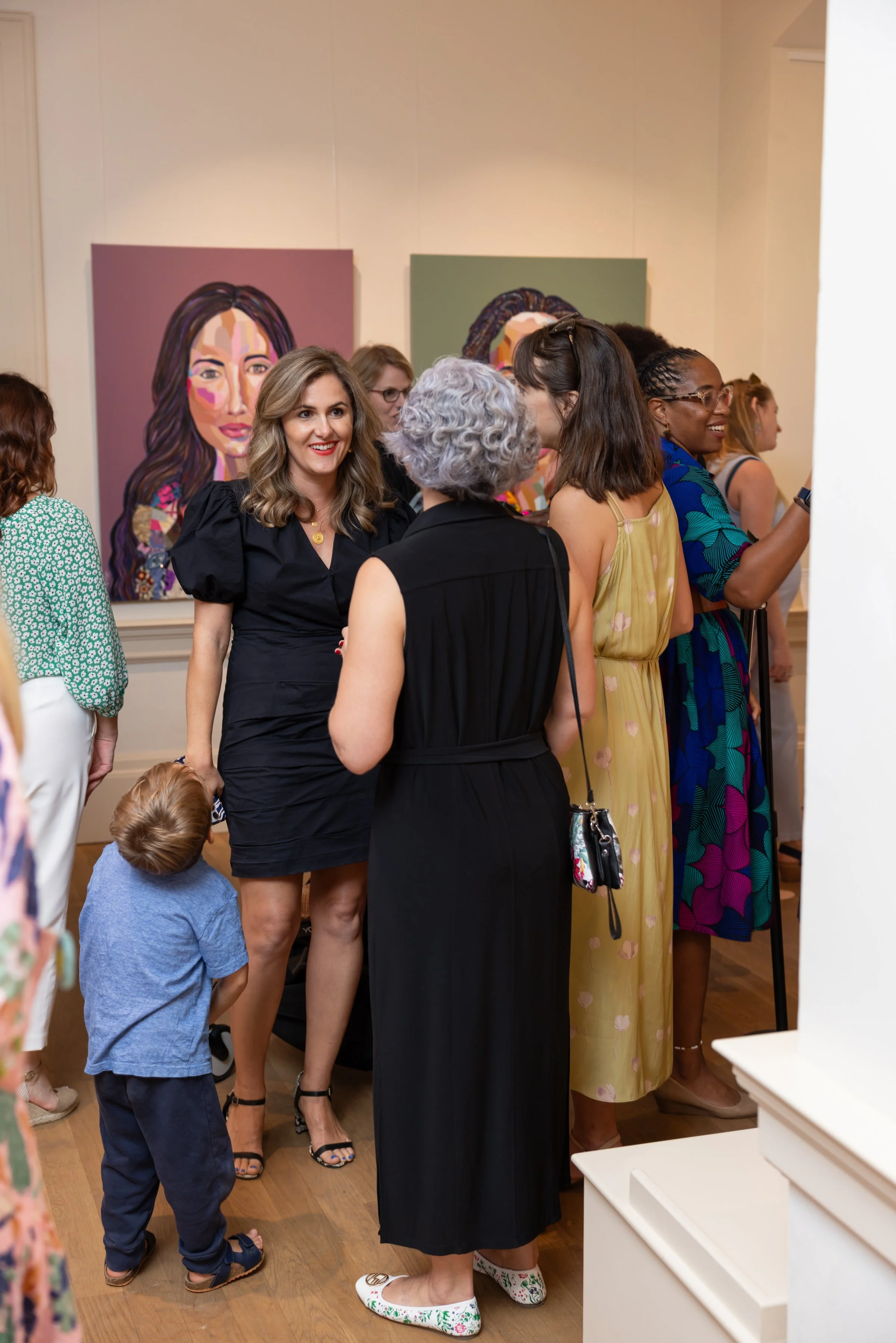 Group of women and children gathered in an art gallery, engaging in conversation and viewing artwork, with colorful portraits of women displayed on the wall in the background.