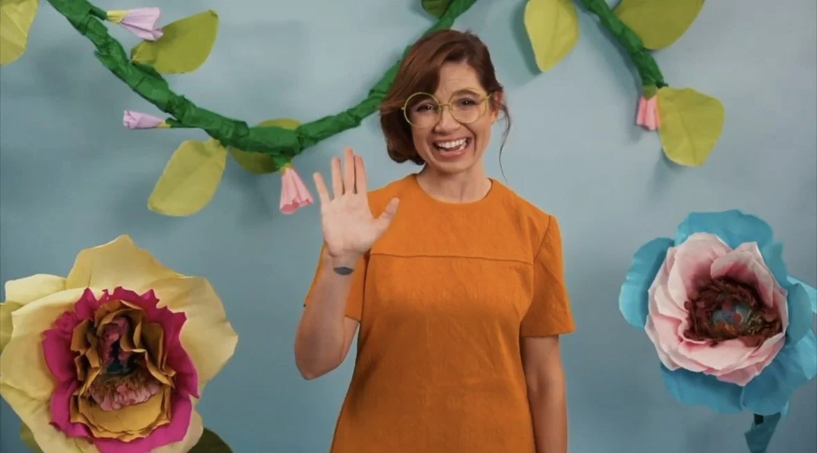 A woman in an orange shirt smiling and waving with large paper flowers and green paper leaves in the background.