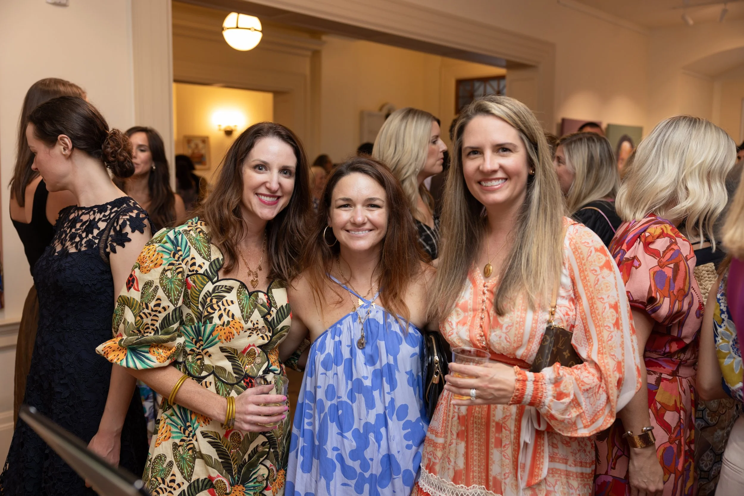 Four women at a social gathering, smiling and holding drinks, indoors with warm lighting and other guests in the background.