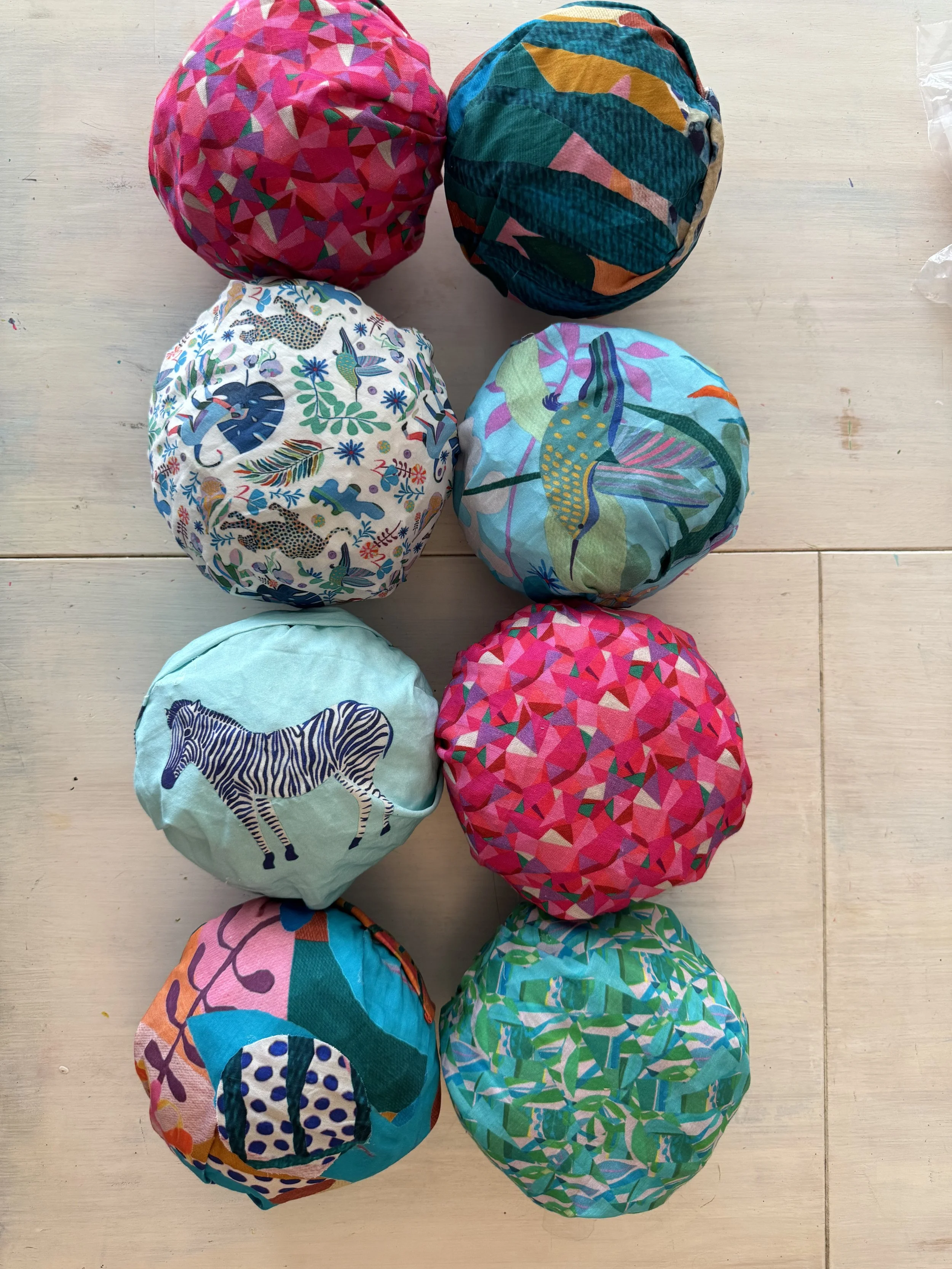 Seven fabric-covered objects, possibly balls or cushions, with colorful, patterned designs, placed on a light wooden surface.
