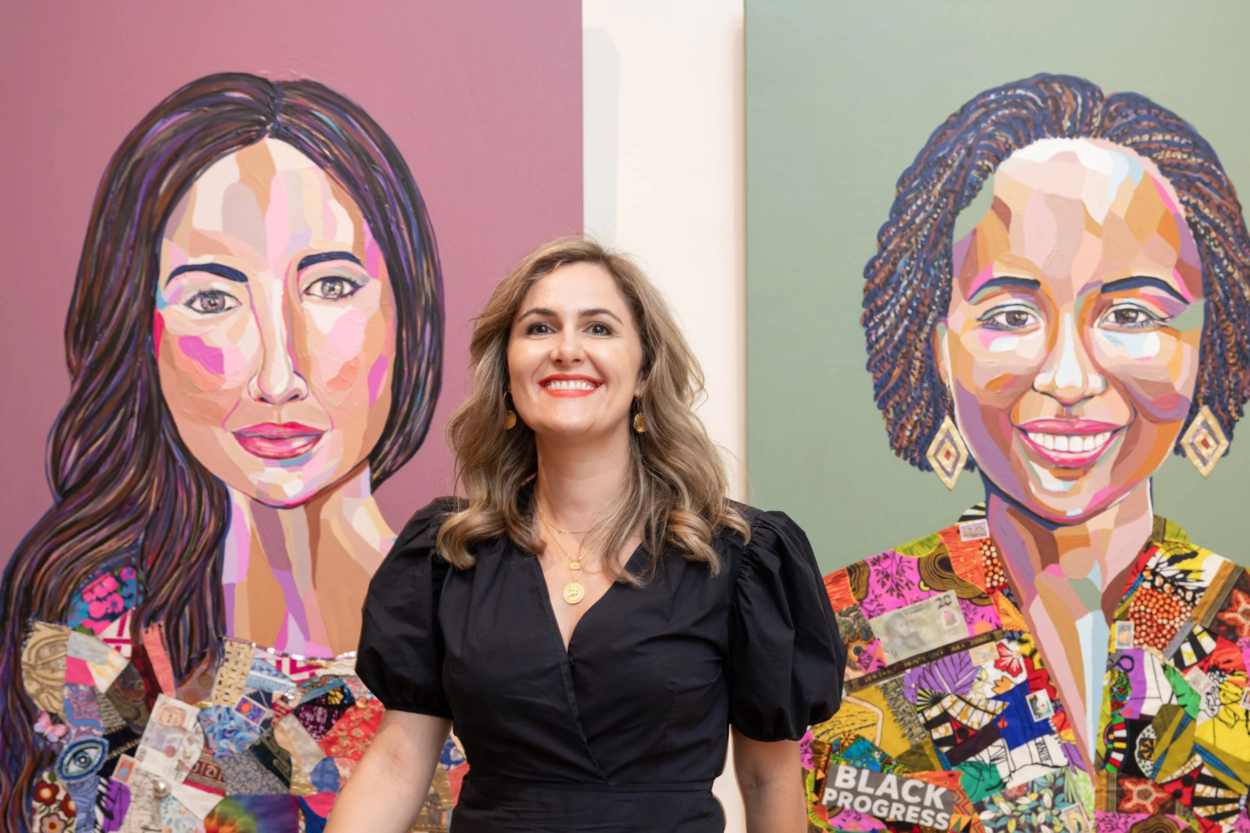 A woman with wavy light brown hair, wearing a black dress and gold jewelry, smiling while standing between two colorful portraits of women with braided hair, vibrant backgrounds, and collage-style clothing.