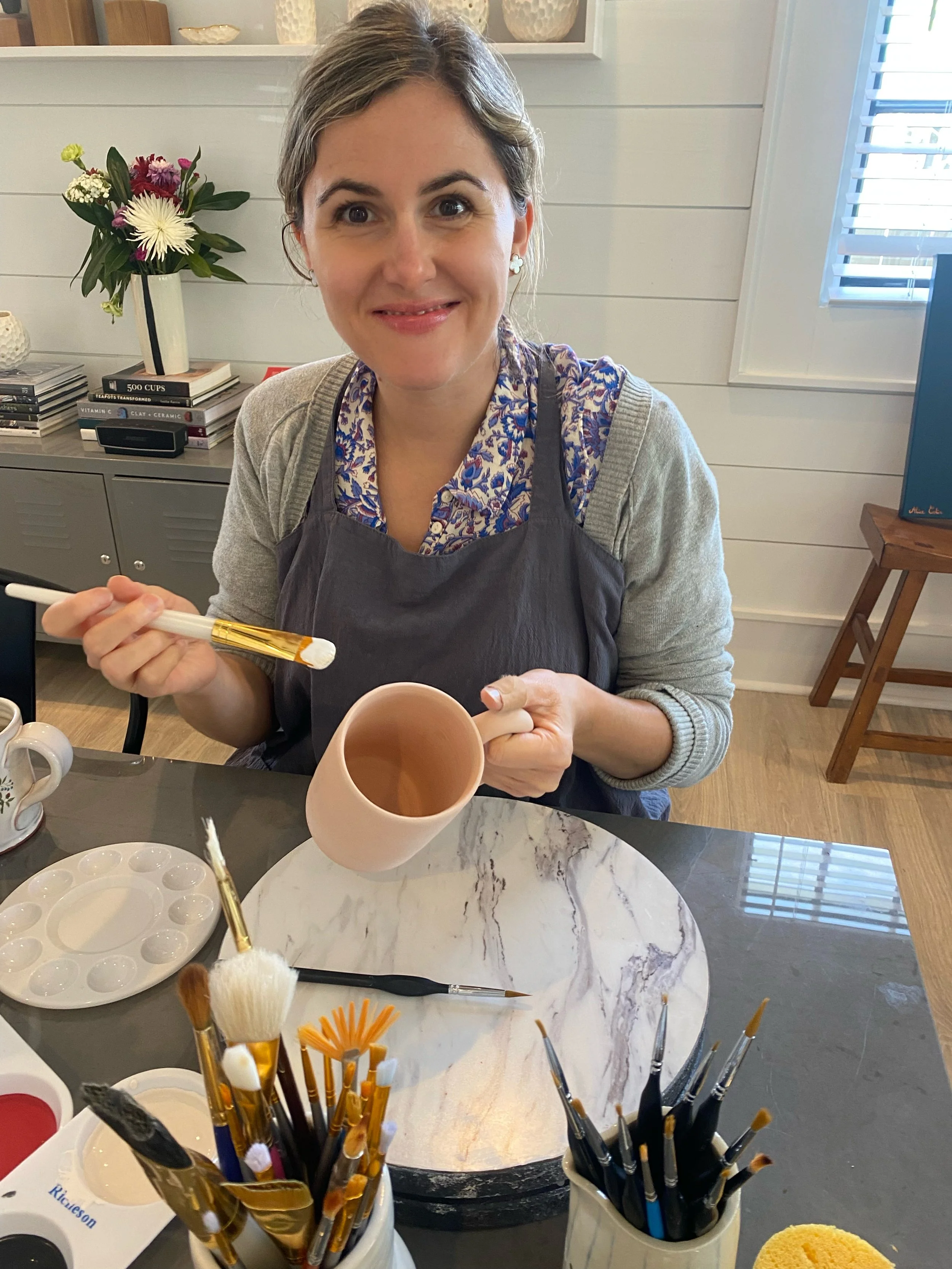 A woman with short brown hair and blue eyes, wearing a gray apron over a blue patterned shirt, is smiling while holding a paintbrush. In front of her on a marble table are painting supplies, including brushes, paint palettes, and a ceramic mug. In the background, there is a vase with flowers and books on a gray cabinet, and a window with blinds.