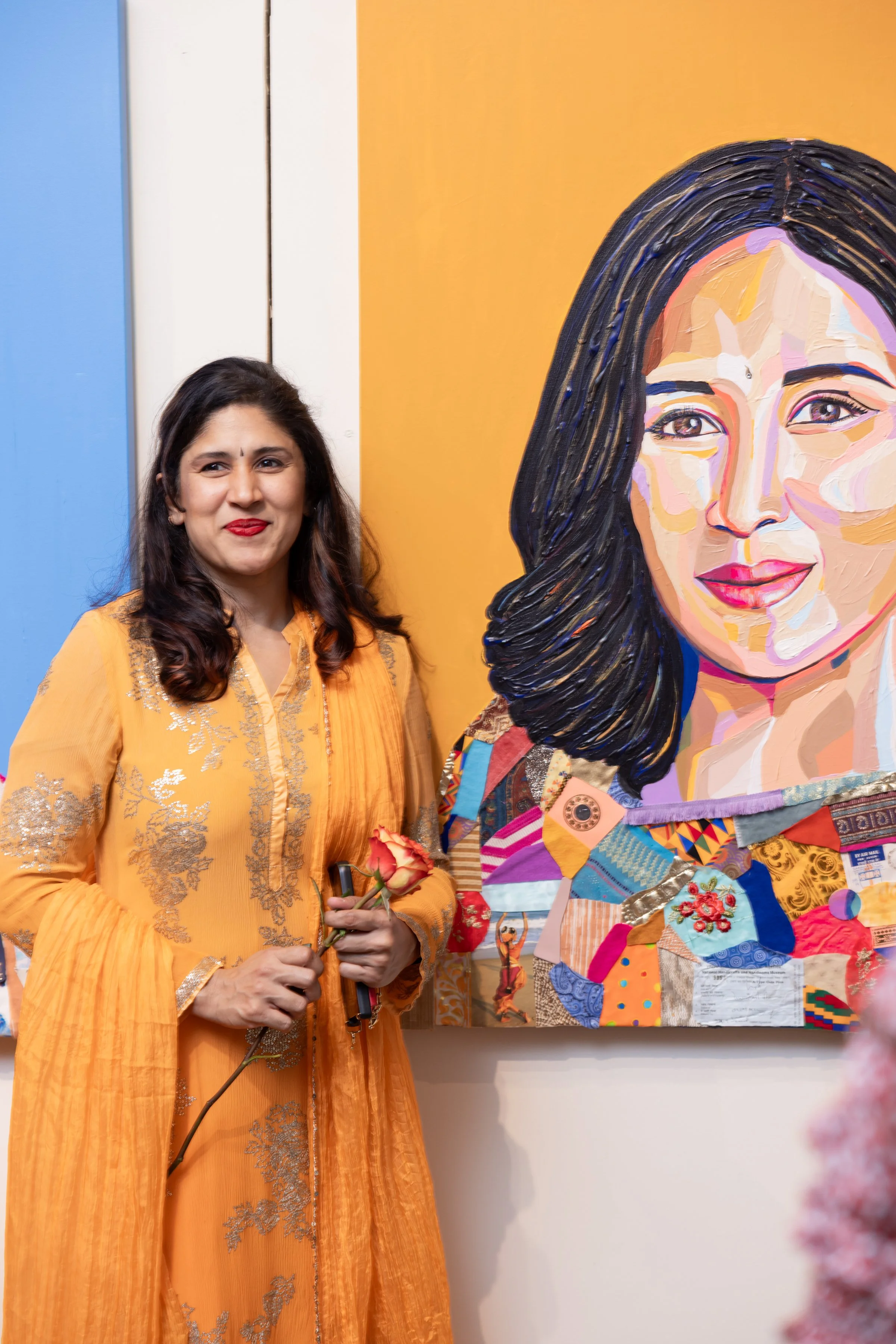 A woman dressed in an orange traditional outfit holding a rose standing next to a large colorful portrait painting of a woman's face.