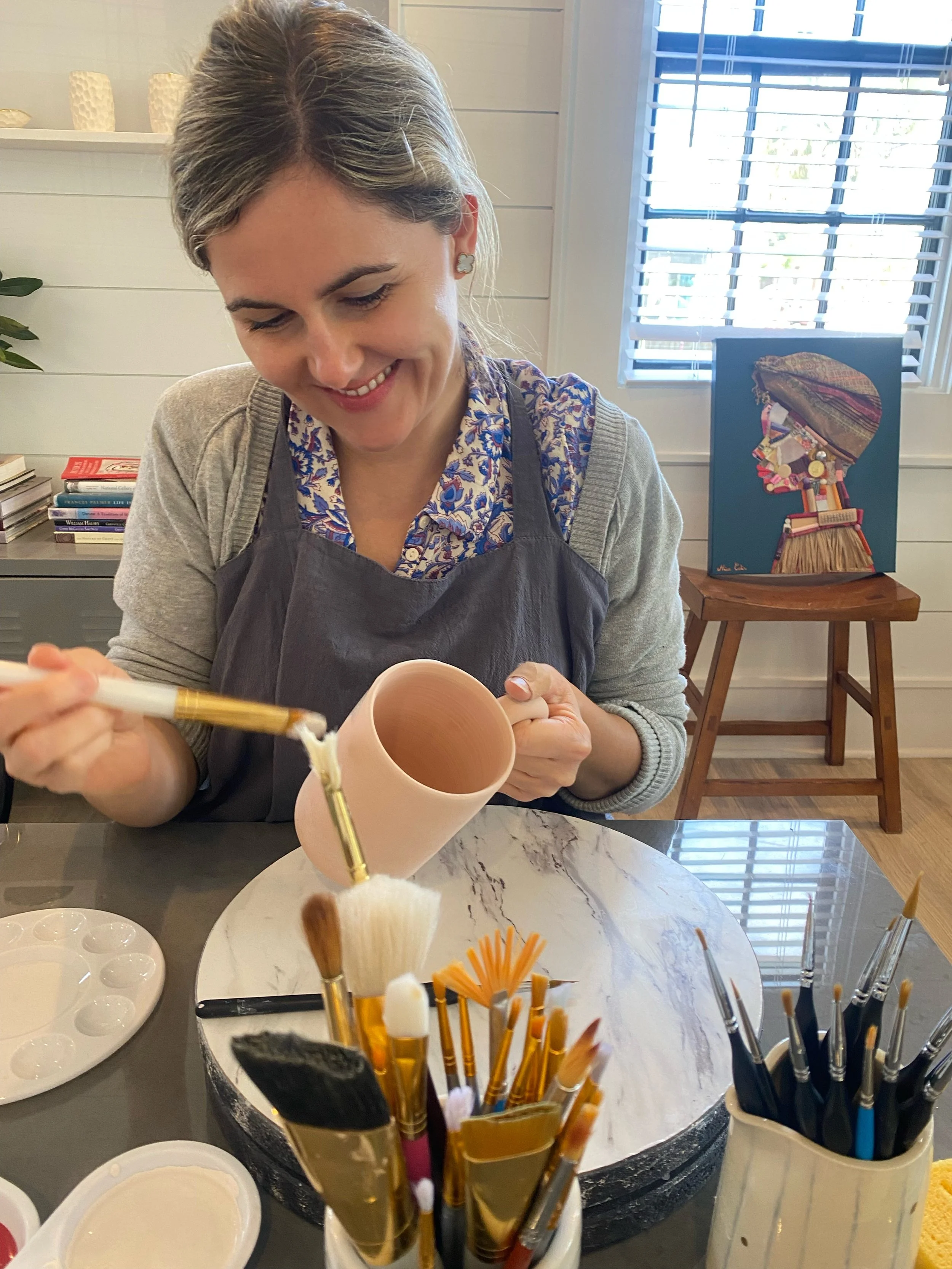 A woman with gray hair smiling and holding a pink ceramic mug, working on an art project in a well-lit indoor space with white shiplap walls and window blinds. Behind her, there is an easel with a colorful mixed media artwork of a person's profile silhouette. The table in front of her holds various art supplies, including brushes, palettes, and containers.