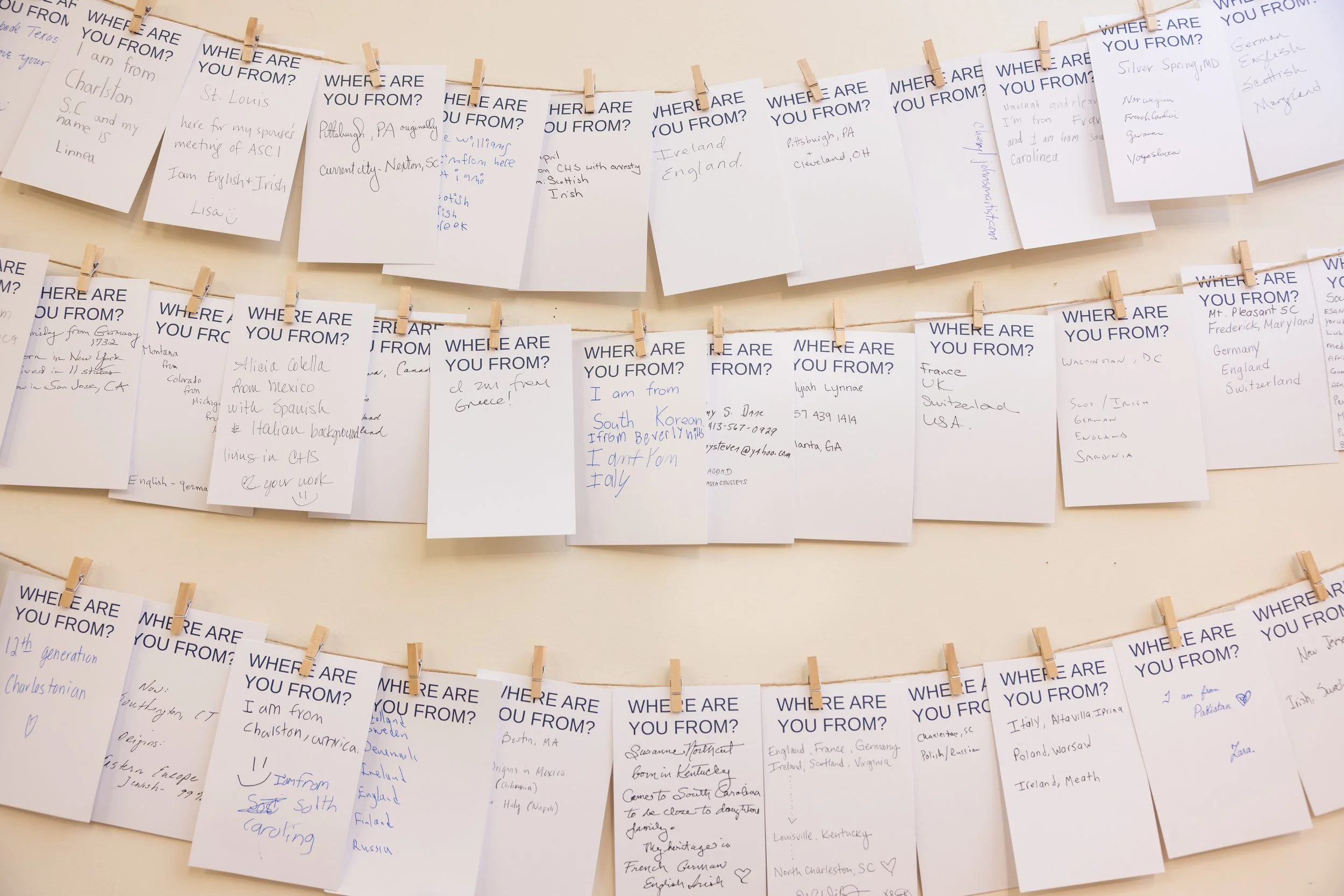 Multiple white cards with handwritten responses to the question, 'Where are you from?', clipped onto strings with small clothespins, arranged on a wall.