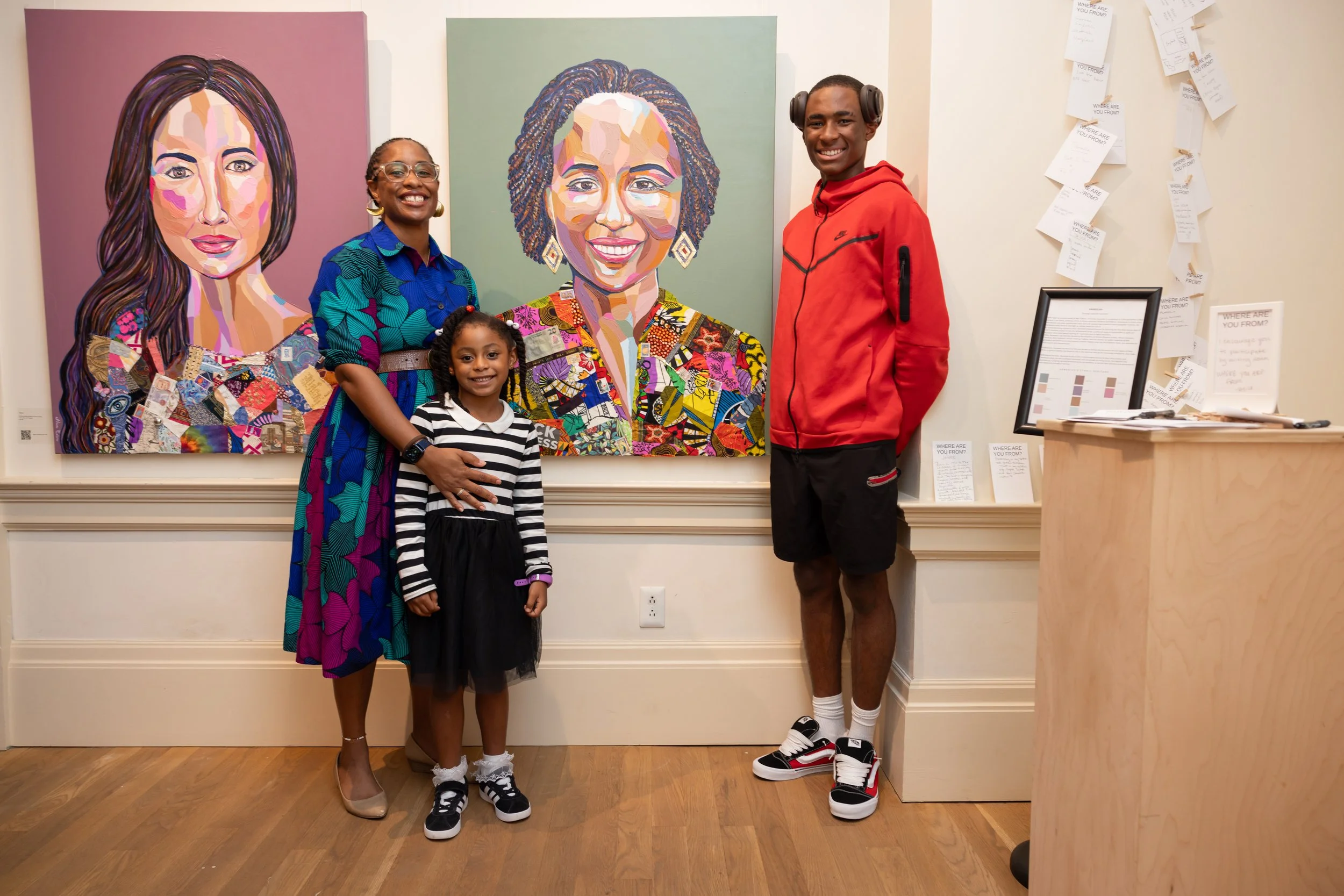 A group of four people standing in front of two large, colorful, portrait paintings of women. The group includes two women, a young girl, and a teenage boy. They are smiling and appear happy. The setting seems to be an art gallery or exhibition space