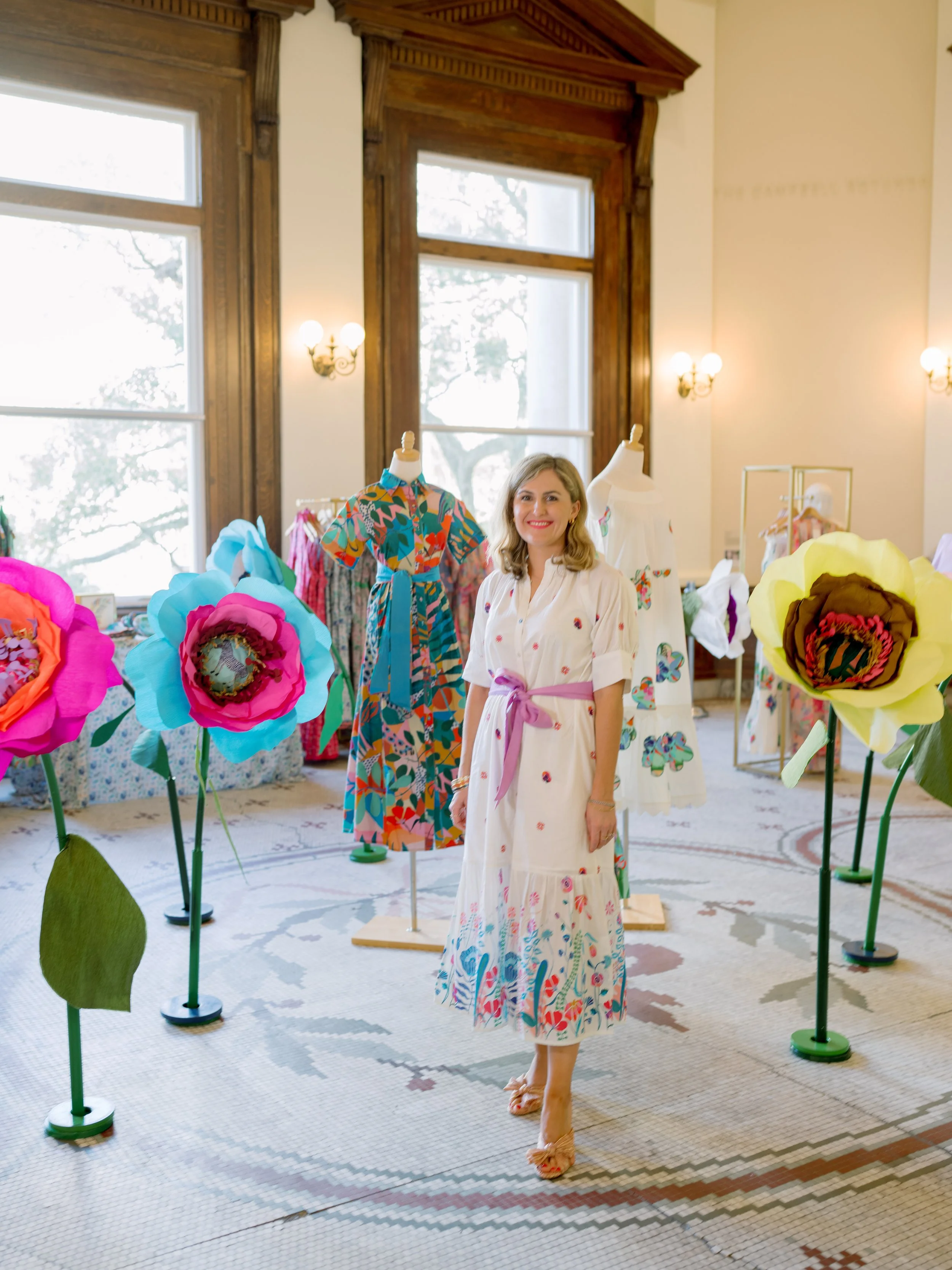 A woman standing in a room decorated with large, colorful paper flowers and mannequins wearing floral dresses. She is smiling and wearing a white dress with butterfly embroidery and a pink sash. The room has tall windows, wooden framing, and vintage 