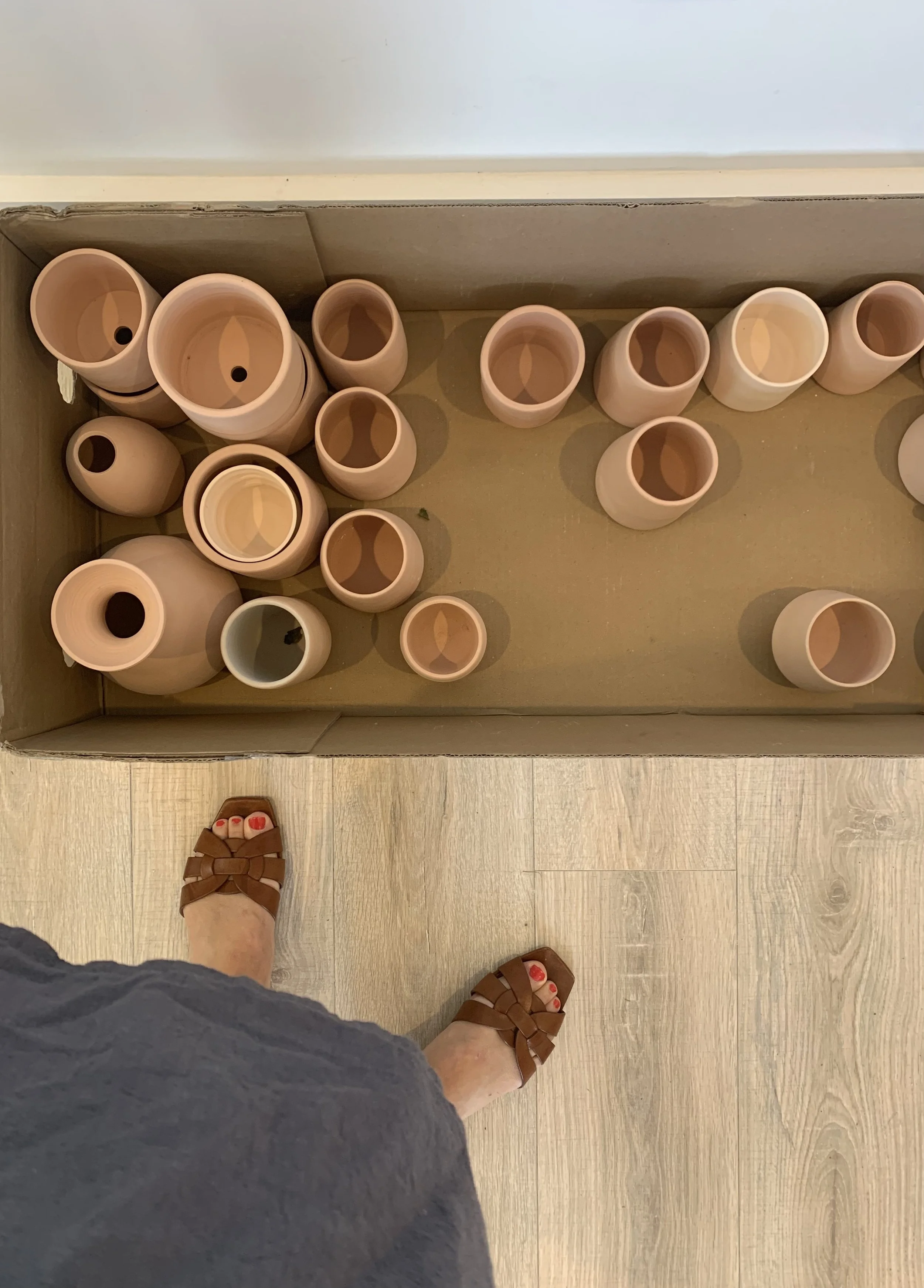 A view of a person standing on a light wood floor, wearing brown woven sandals with red painted toenails, looking down at a cardboard box filled with empty beige ceramic vases.