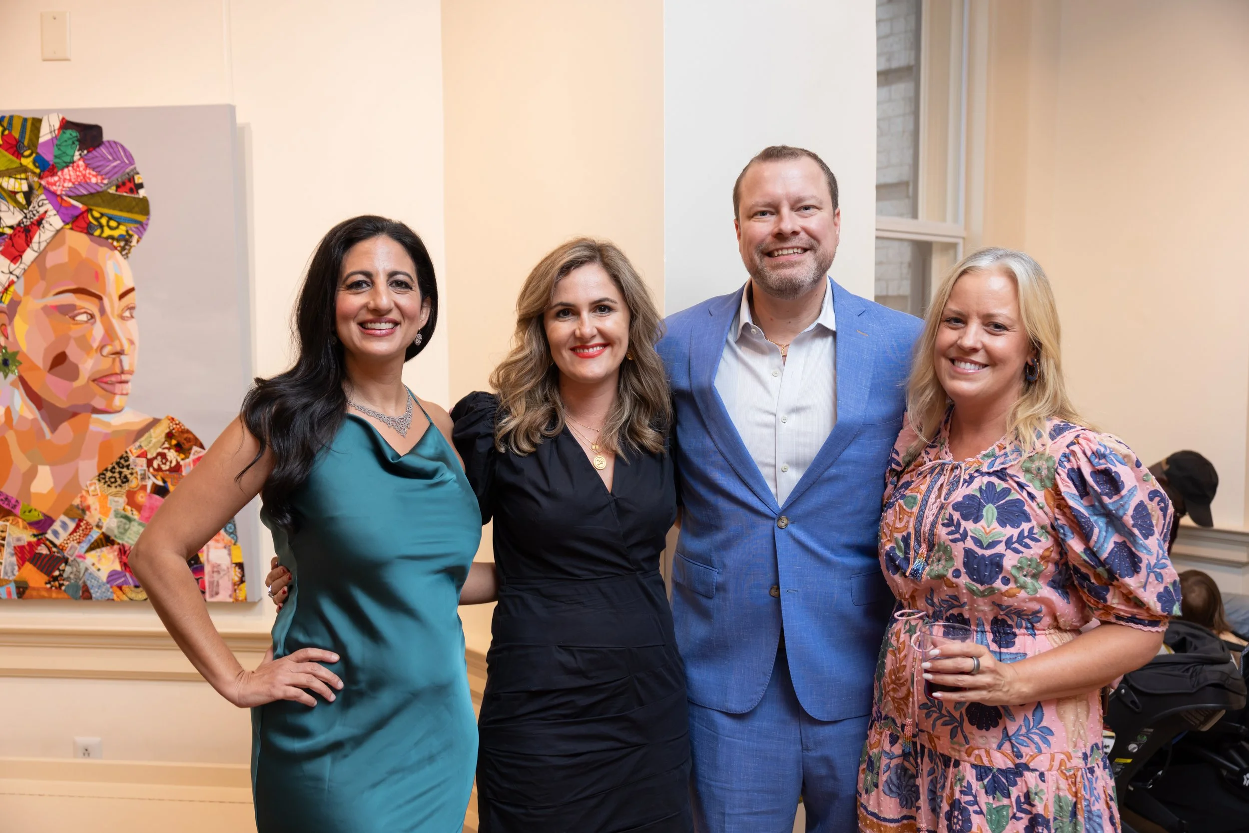 Group of four smiling adults, three women and one man, standing inside a well-lit room with white walls and large windows, dressed in colorful and professional attire, with an abstract portrait artwork visible in the background.