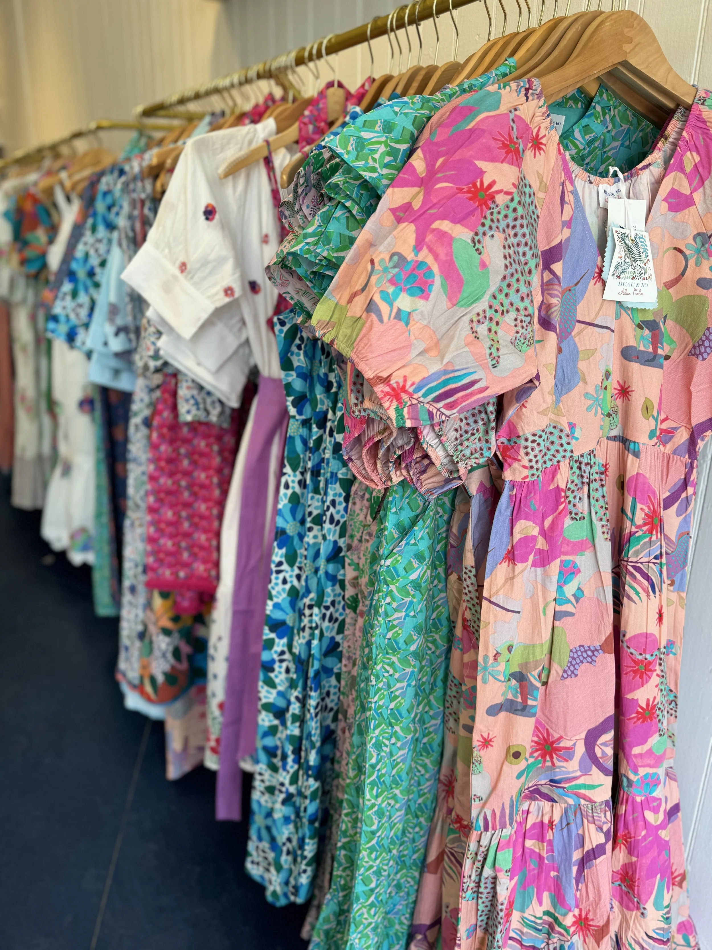 A rack of colorful summer dresses and clothing hanging on wooden hangers, with various patterns including floral and abstract designs in shades of pink, blue, green, and purple.