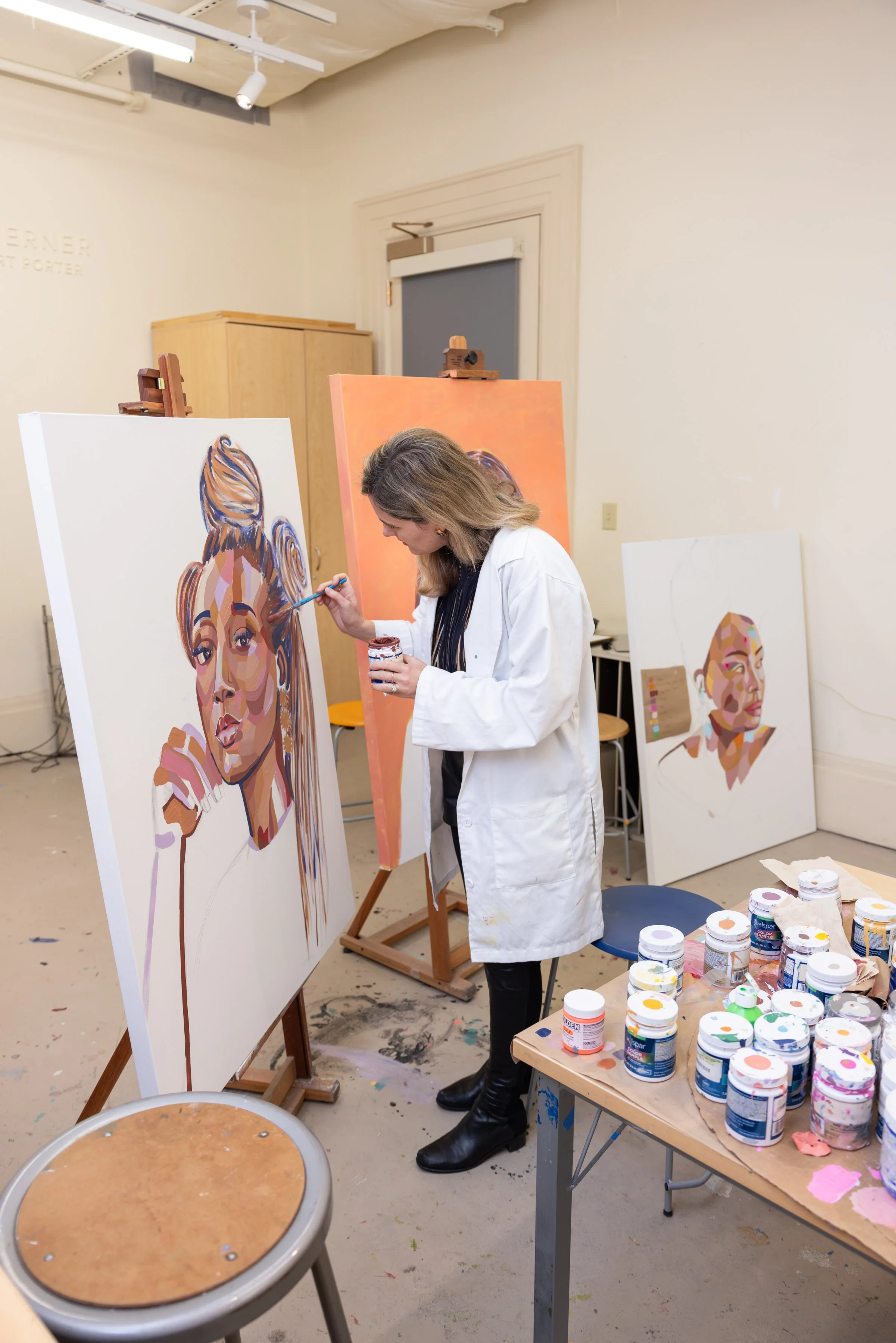 An artist wearing a white coat is painting a colorful portrait of a woman on a large canvas. The studio has multiple paintings, art supplies, and a wooden cabinet in the background.