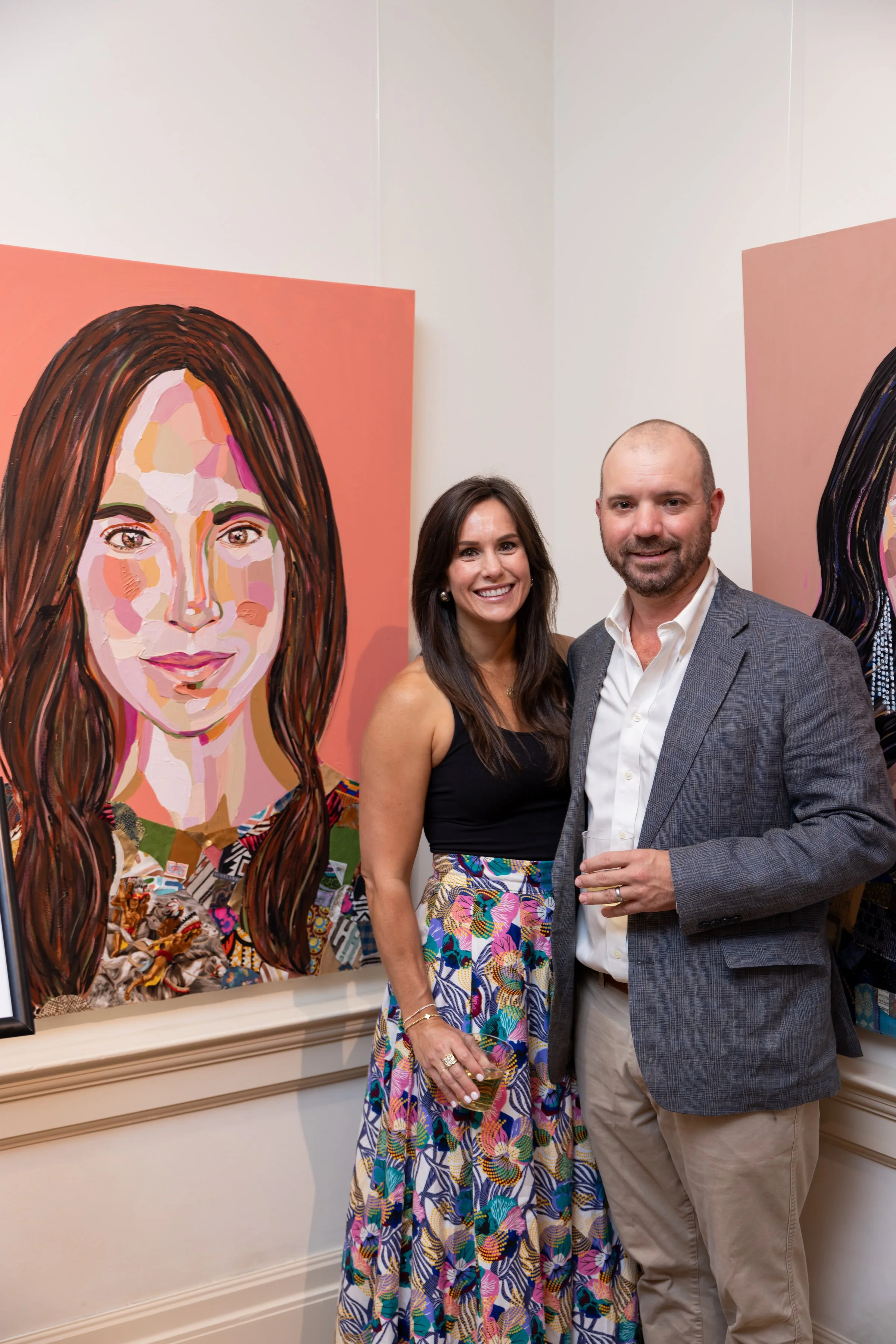 A woman and a man stand together at an art gallery, smiling. The woman is wearing a black top and a colorful patterned skirt, holding a drink. The man is wearing a gray blazer and beige pants, holding a glass. Behind them are two large colorful abstr