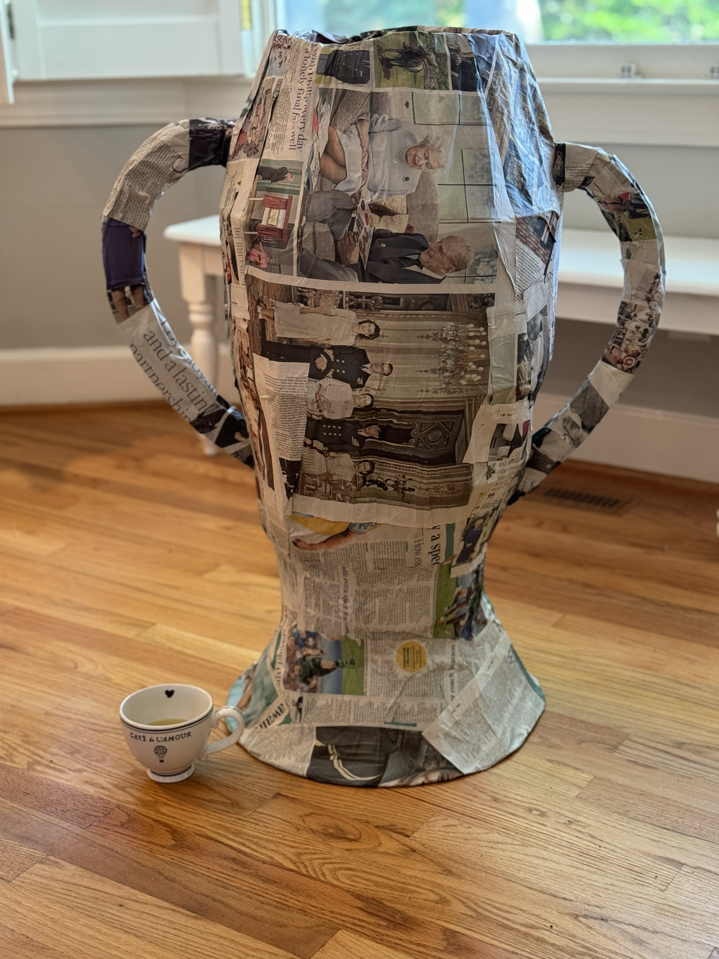 A paper mache sculpture of a woman with a dress and a headscarf, made from newspaper clippings, placed on a wooden table with a white mug nearby. The background features a window and a gray wall.
