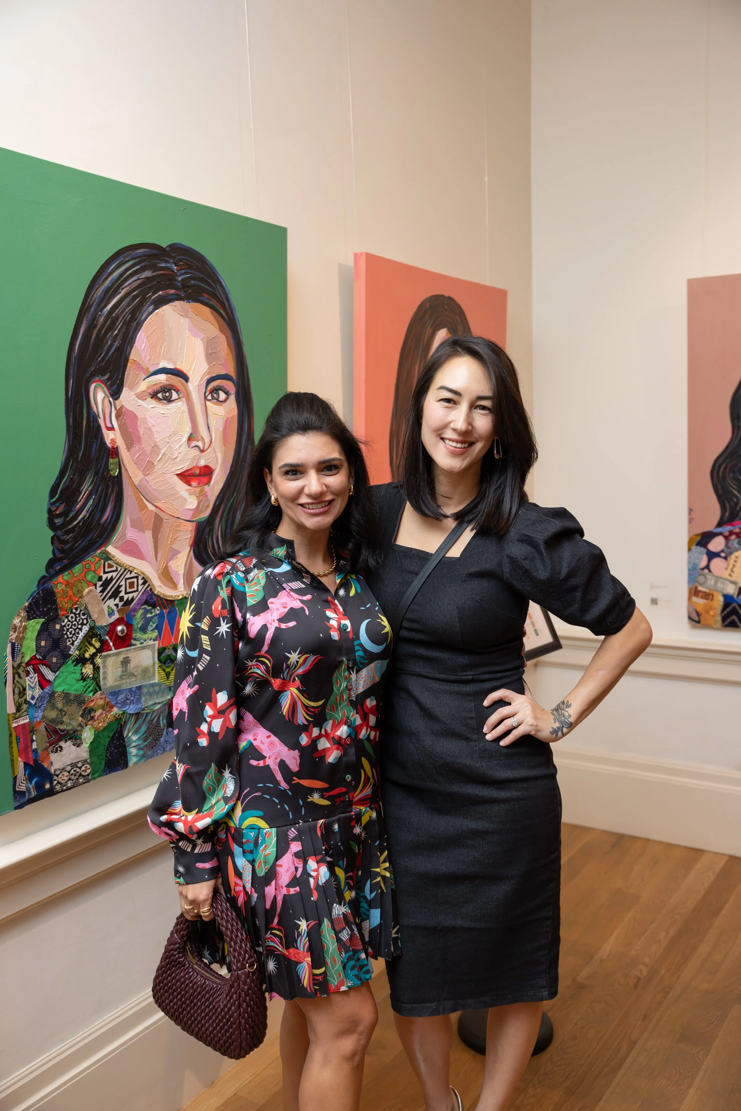 Two women posing in front of colorful portrait paintings at an art gallery. One woman is wearing a colorful patterned dress and holding a maroon handbag, and the other is wearing a black dress with puffed sleeves.