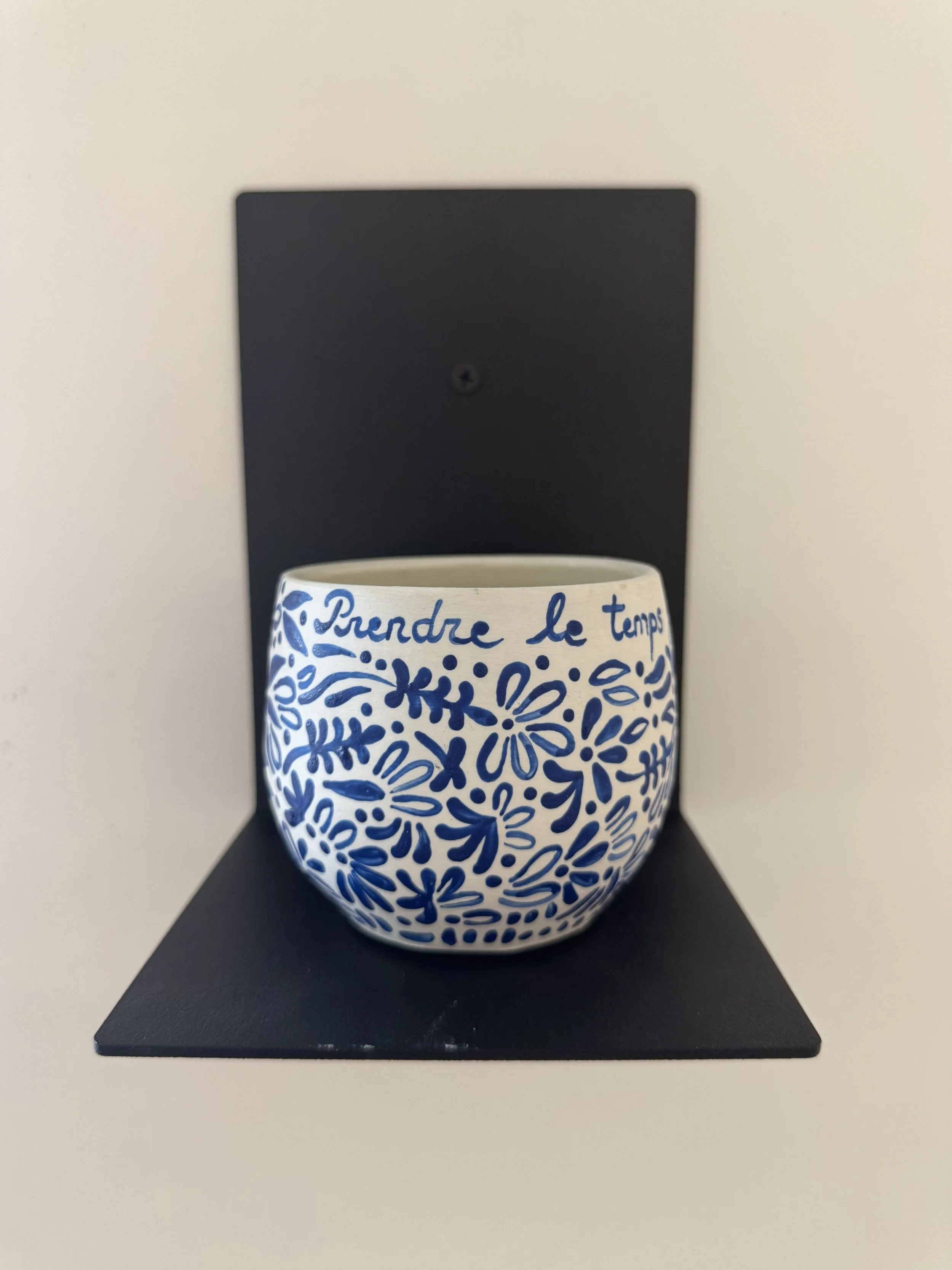 A white ceramic mug with blue floral and text design, placed on a black stand against a beige wall.