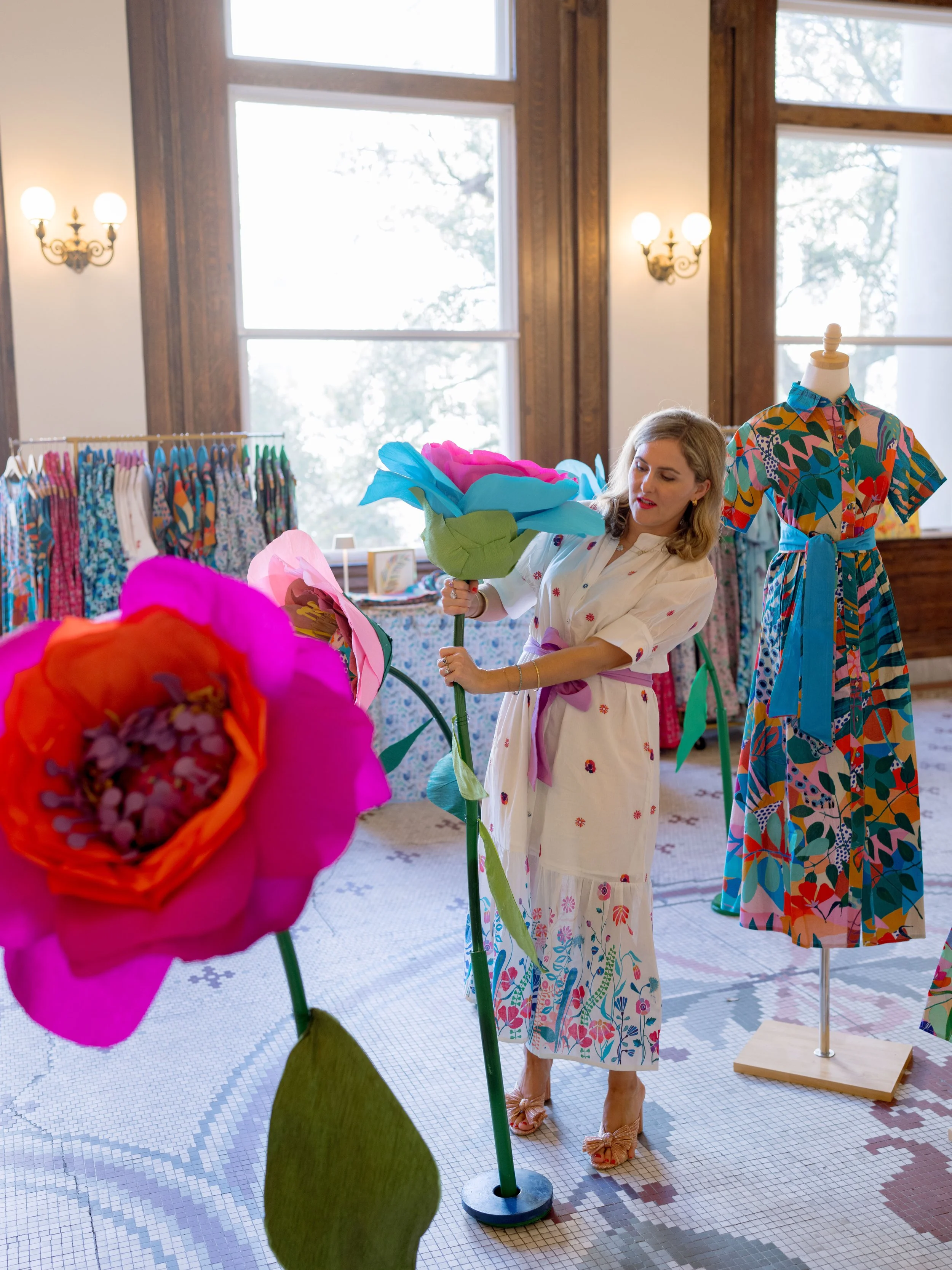 A woman in a floral dress holds a large paper flower in a room with large windows and vintage wall sconces, surrounded by clothing on racks and a dress form.