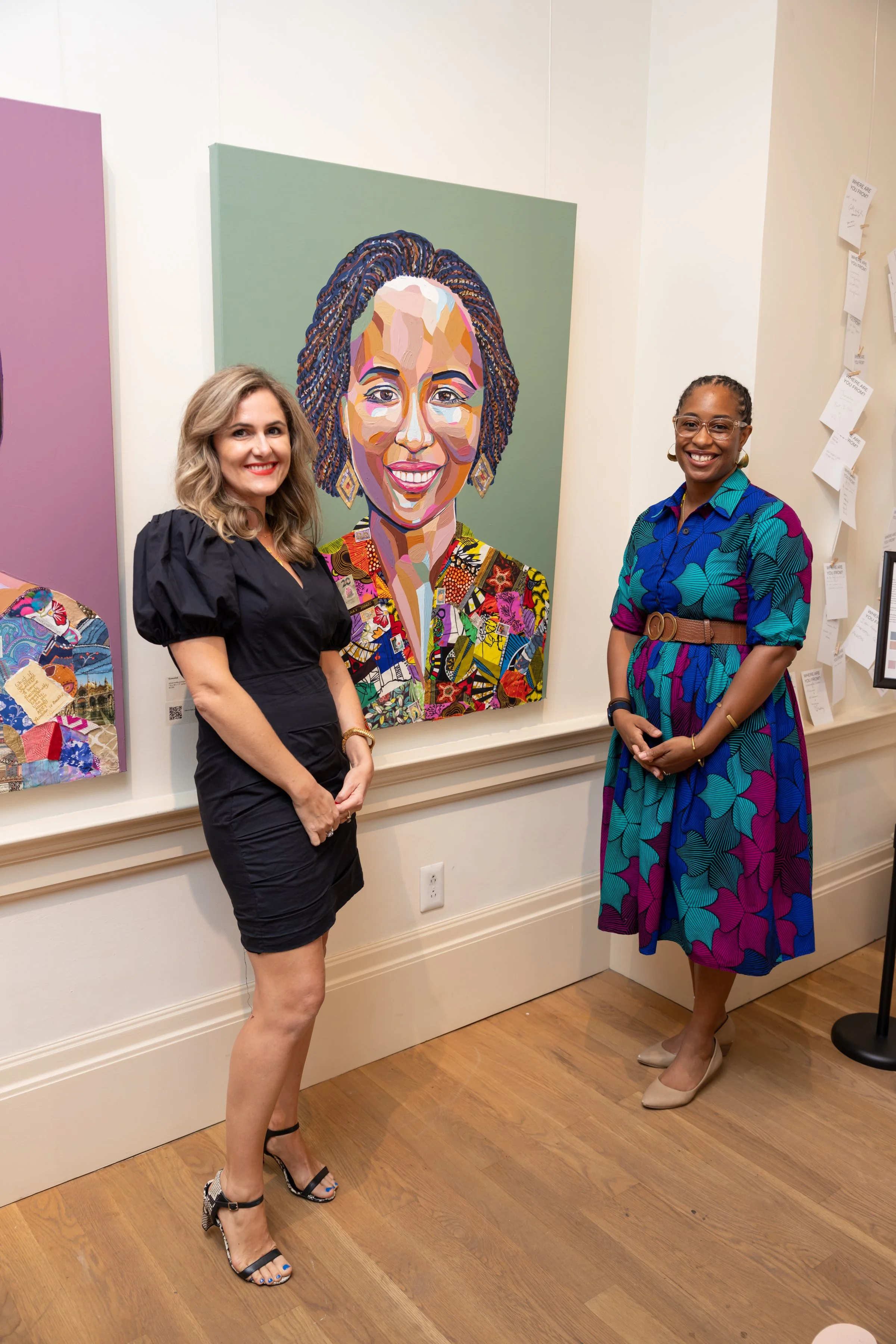Two women standing in front of a colorful portrait painting at an art gallery. The woman on the left is wearing a black dress with puffed sleeves, patterned high heels, and has wavy blonde hair. The woman on the right is wearing a vibrant, patterned 