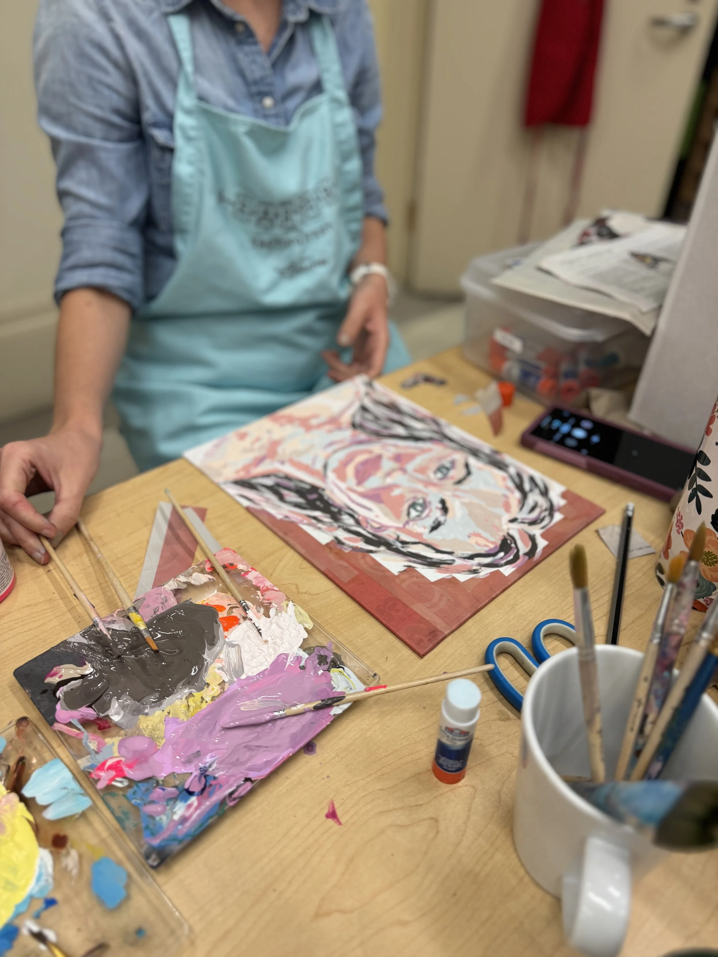 A woman with gray hair smiling and holding a pink ceramic mug, working on an art project in a well-lit indoor space with white shiplap walls and window blinds. Behind her, there is an easel with a colorful mixed media artwork of a person's profile silhouette. The table in front of her holds various art supplies, including brushes, palettes, and containers.
