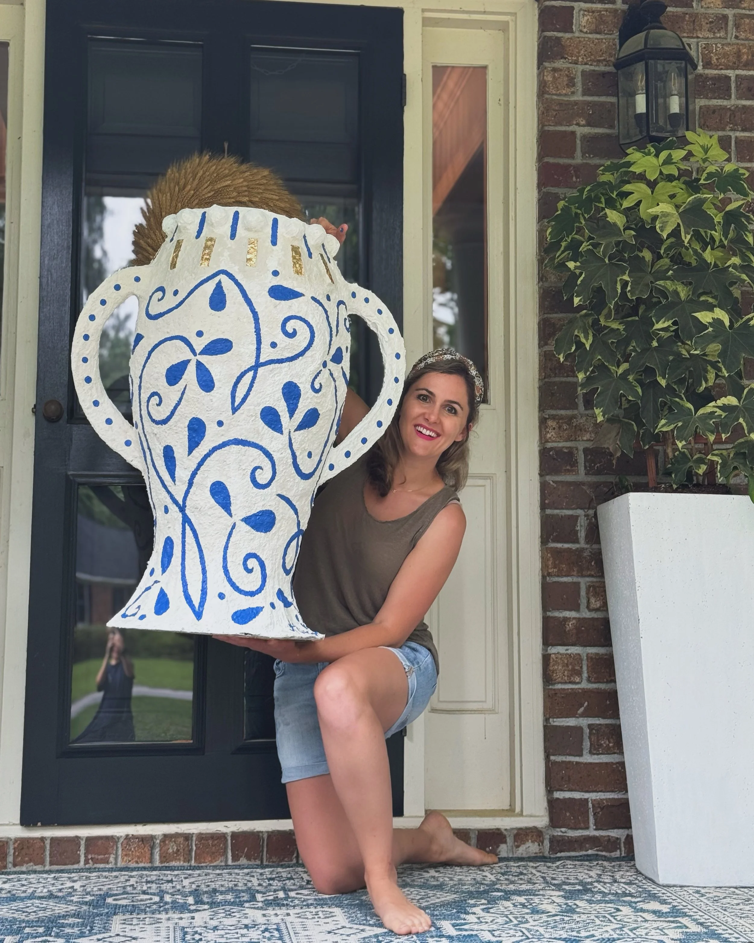 A woman kneeling on a porch steps holding a large decorative white lantern with blue designs, with a smiling face, in front of a black front door with a window. There is a brick wall on the right, a potted ivy plant, and a white outdoor light fixture