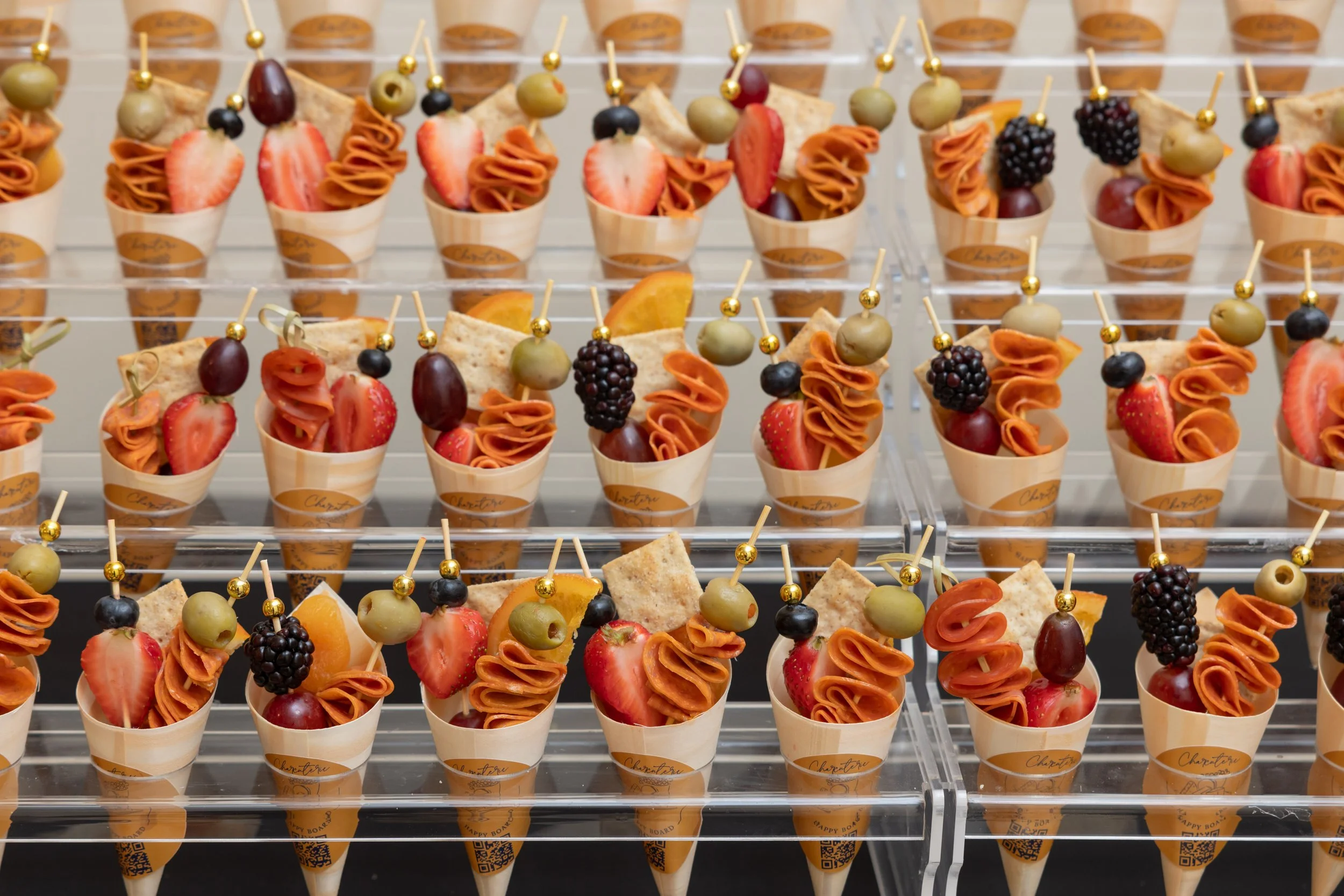 Assorted fruit skewers in small paper cones, arranged on a clear display stand. The skewers include strawberries, grapes, blackberries, and slices of melon with some rolled orange-colored vegetable or fruit. Each cone has a gold sticker with writing 