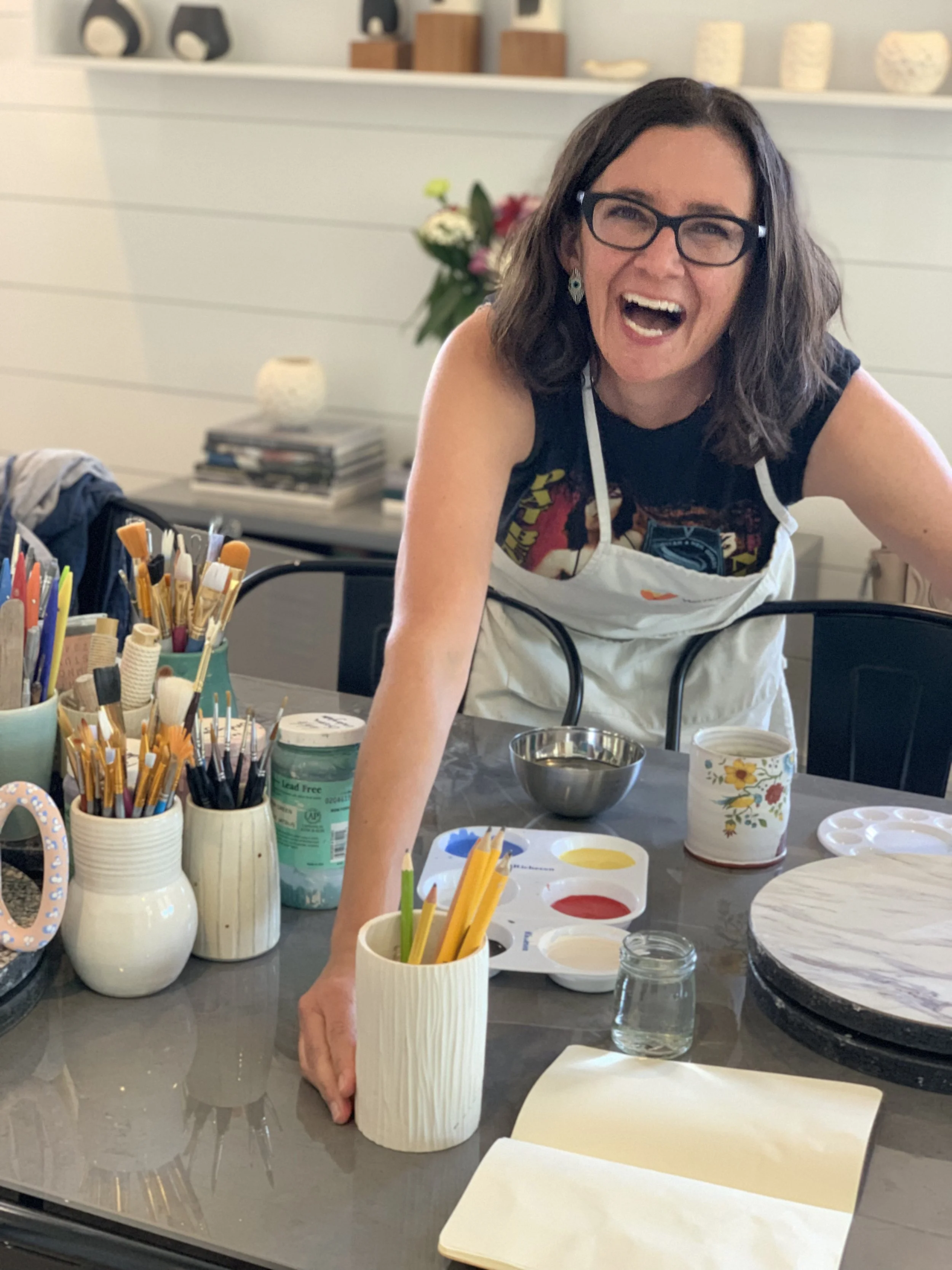 Woman with glasses and dark hair smiling, engaged in a painting or craft activity at a table. The table holds various painting supplies, including brushes, paints, a palette, jars, and paper. Background has decorative items and a bouquet of flowers.
