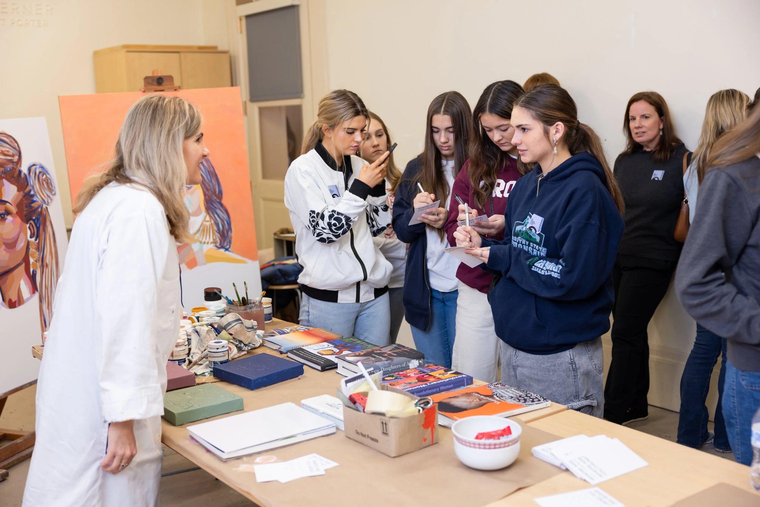 A group of people at an indoor event, looking at a table with books, art supplies, and papers, with artwork displayed in the background.