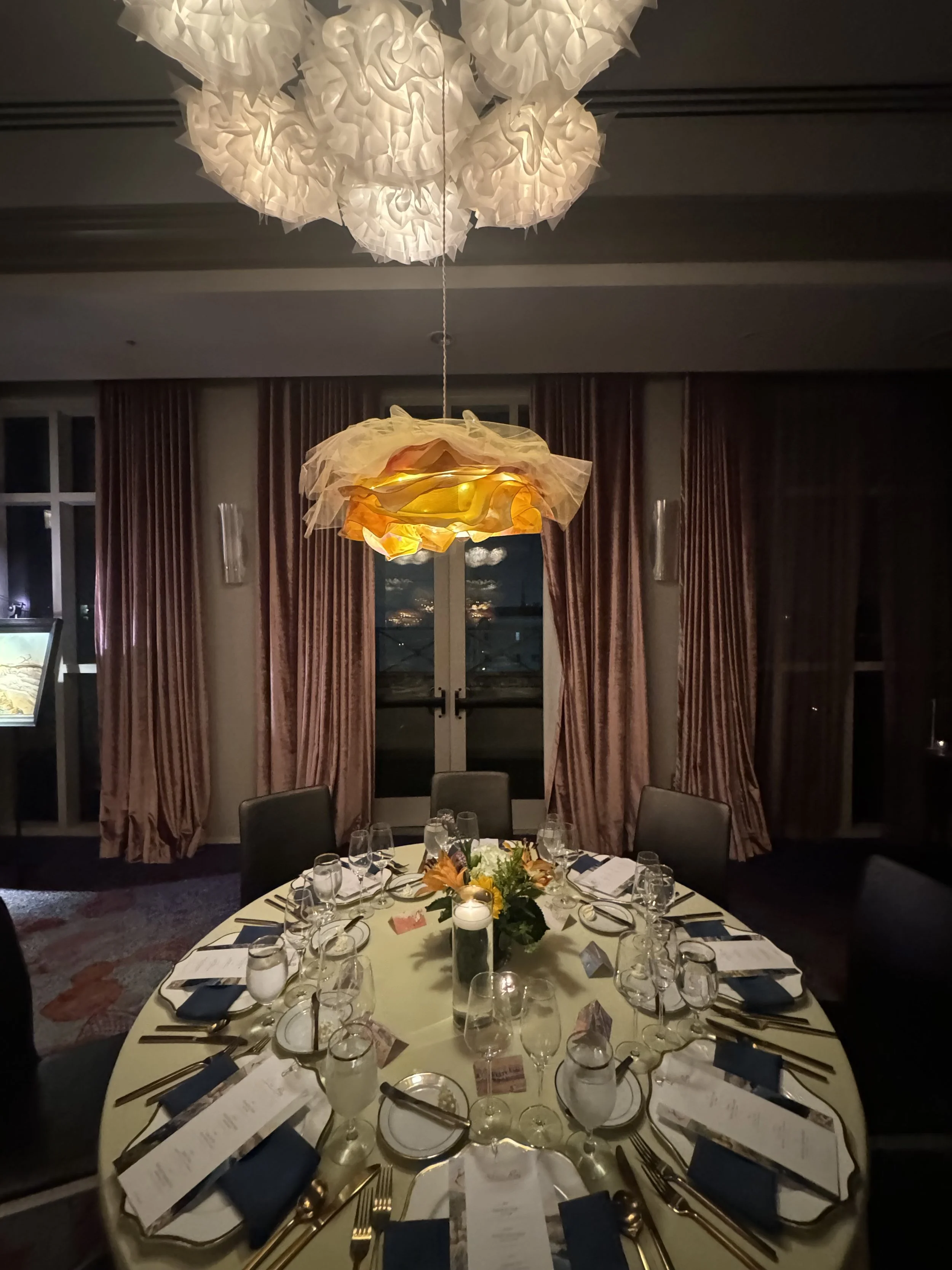 A round dinner table set for a formal event with white tablecloth, wine glasses, silverware, navy blue napkins, menu cards, and a floral centerpiece with yellow flowers and greenery. The room is softly lit with overhead lights and has large windows with pinkish curtains.