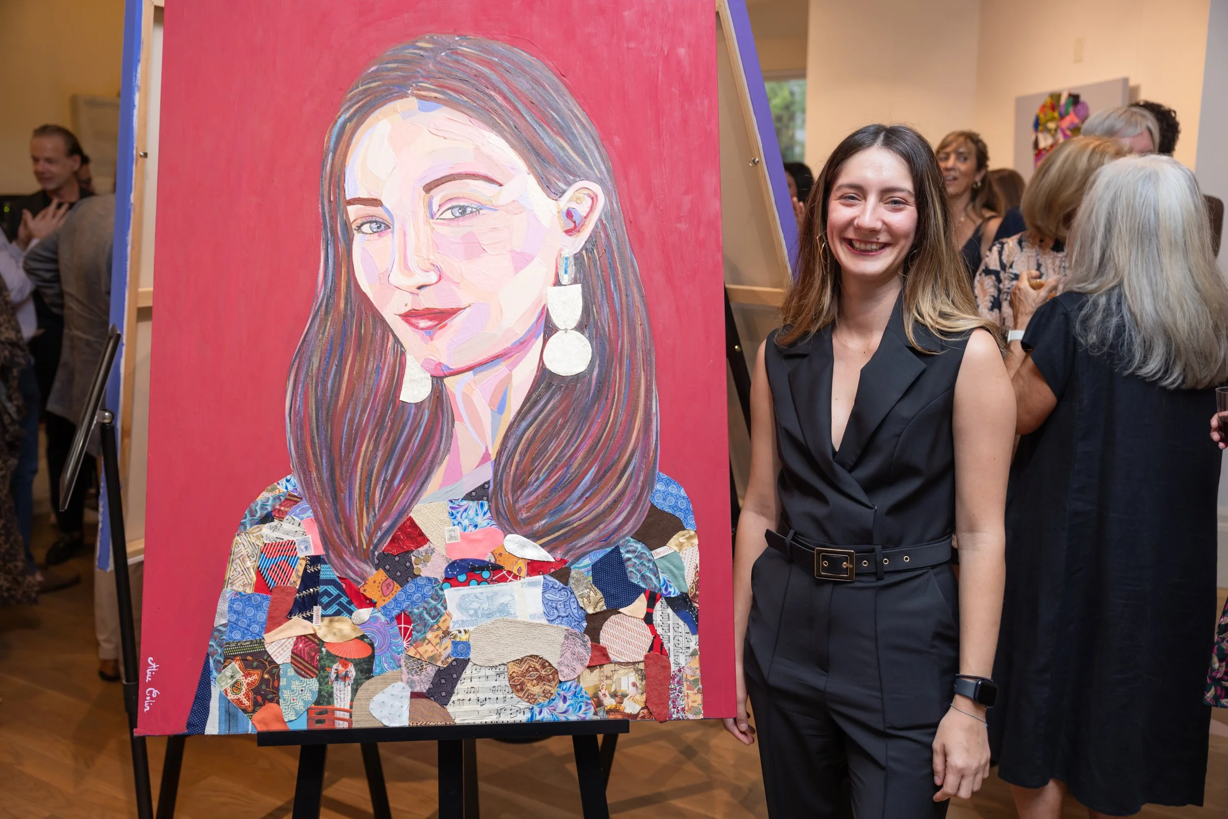 A woman with long brown hair and a black sleeveless outfit smiling while standing next to a colorful abstract portrait painting of a woman with long hair, earrings, and wearing a patterned top, at an art gallery event with people in the background.