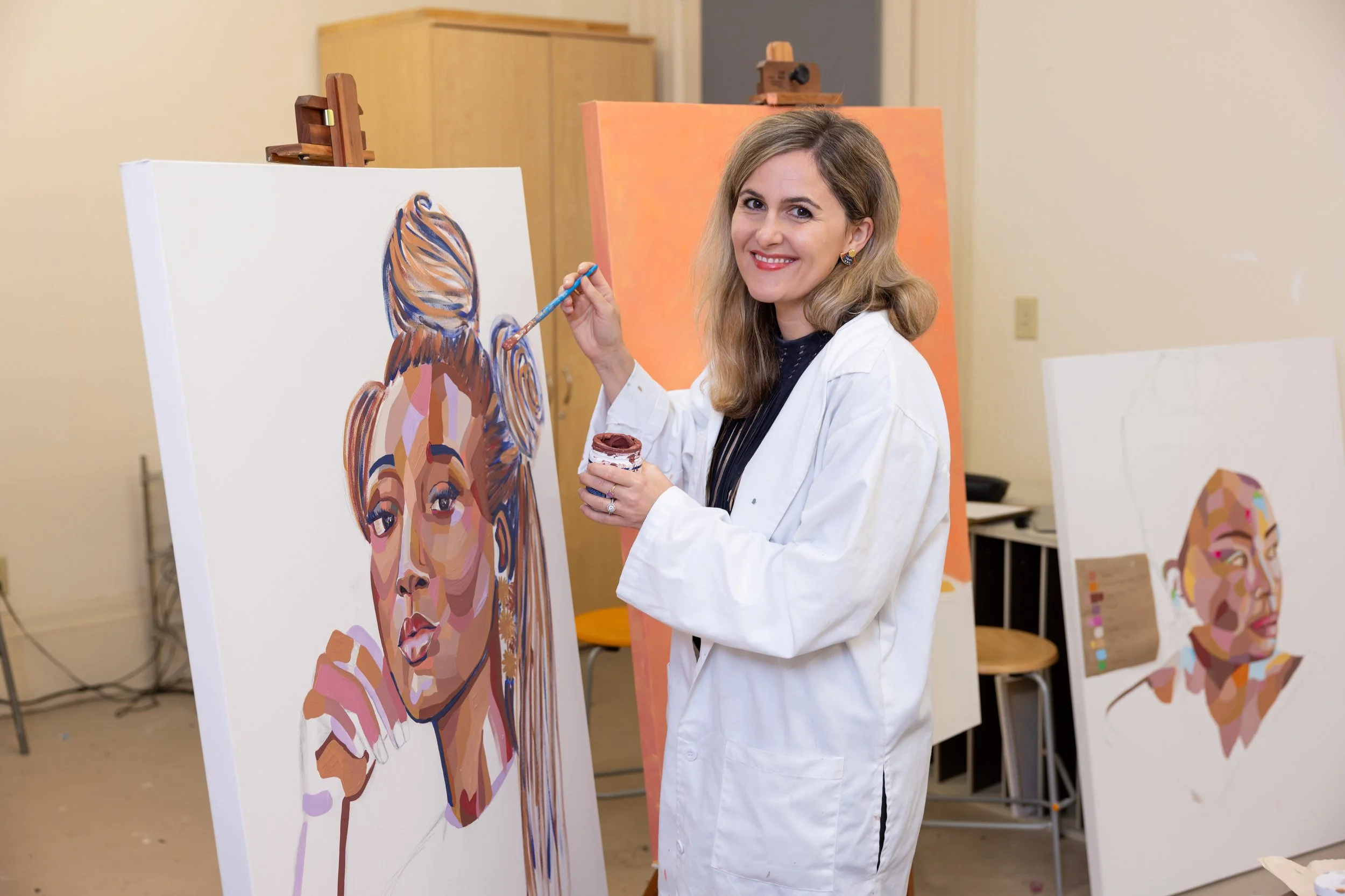 A woman in a white paint smock is painting a colorful, geometric portrait of a woman's face on a canvas in an art studio. She has shoulder-length blonde hair and is smiling while holding a paintbrush and a small container of paint.
