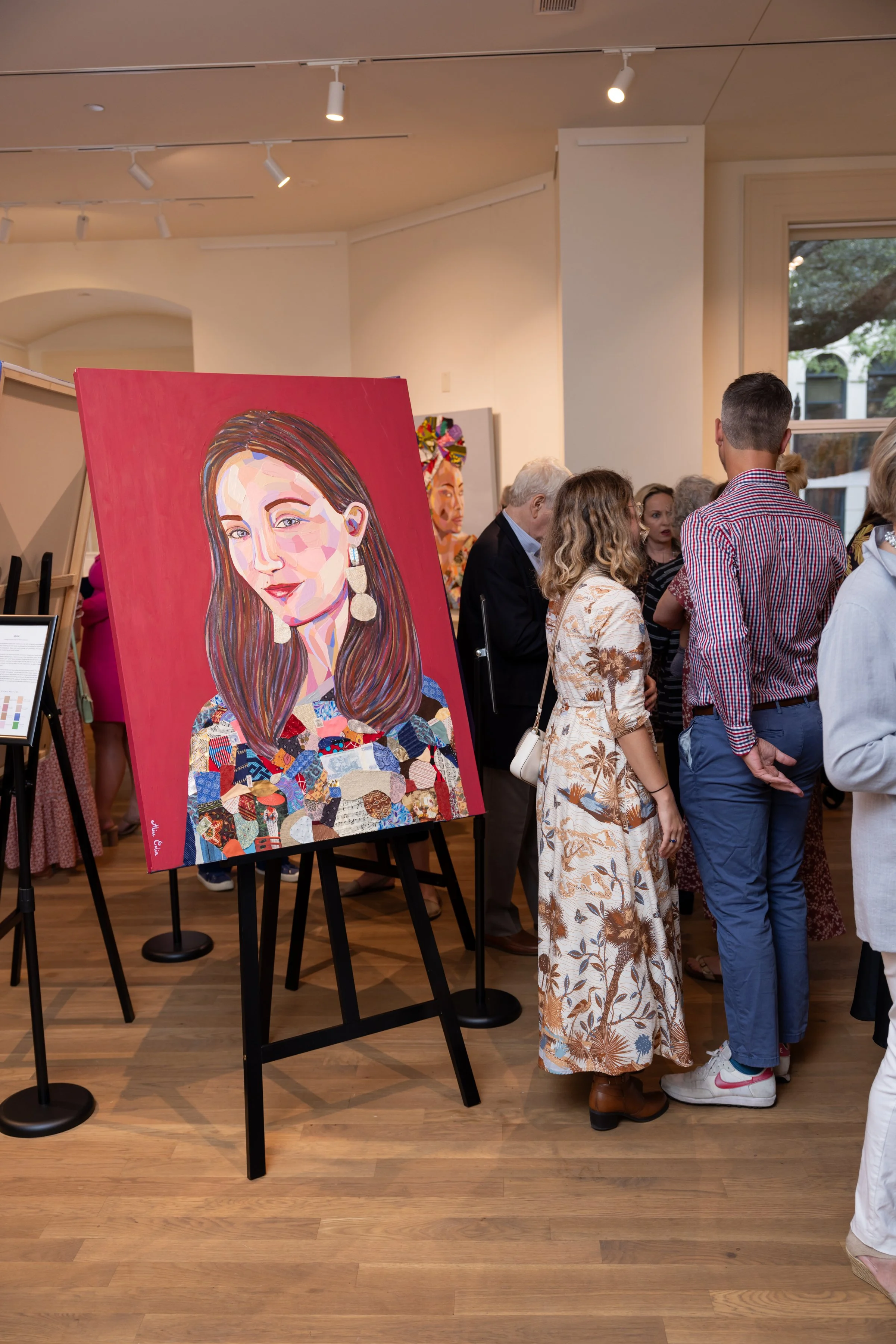 An art gallery with a colorful portrait painting of a woman with long brown hair and earrings on an easel. Several people are gathered and observing artwork, some engaged in conversation.