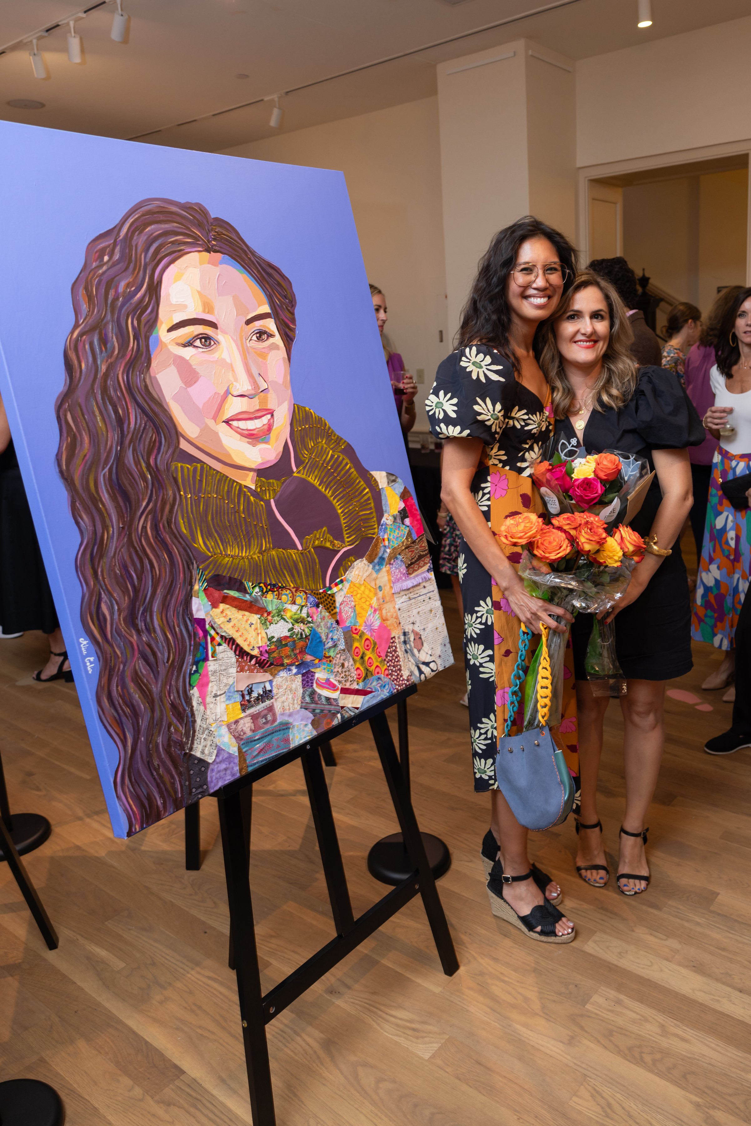 Two women smiling, standing indoors at an art exhibition with a large colorful portrait of a woman on an easel in the foreground. One woman holds a bouquet of flowers.