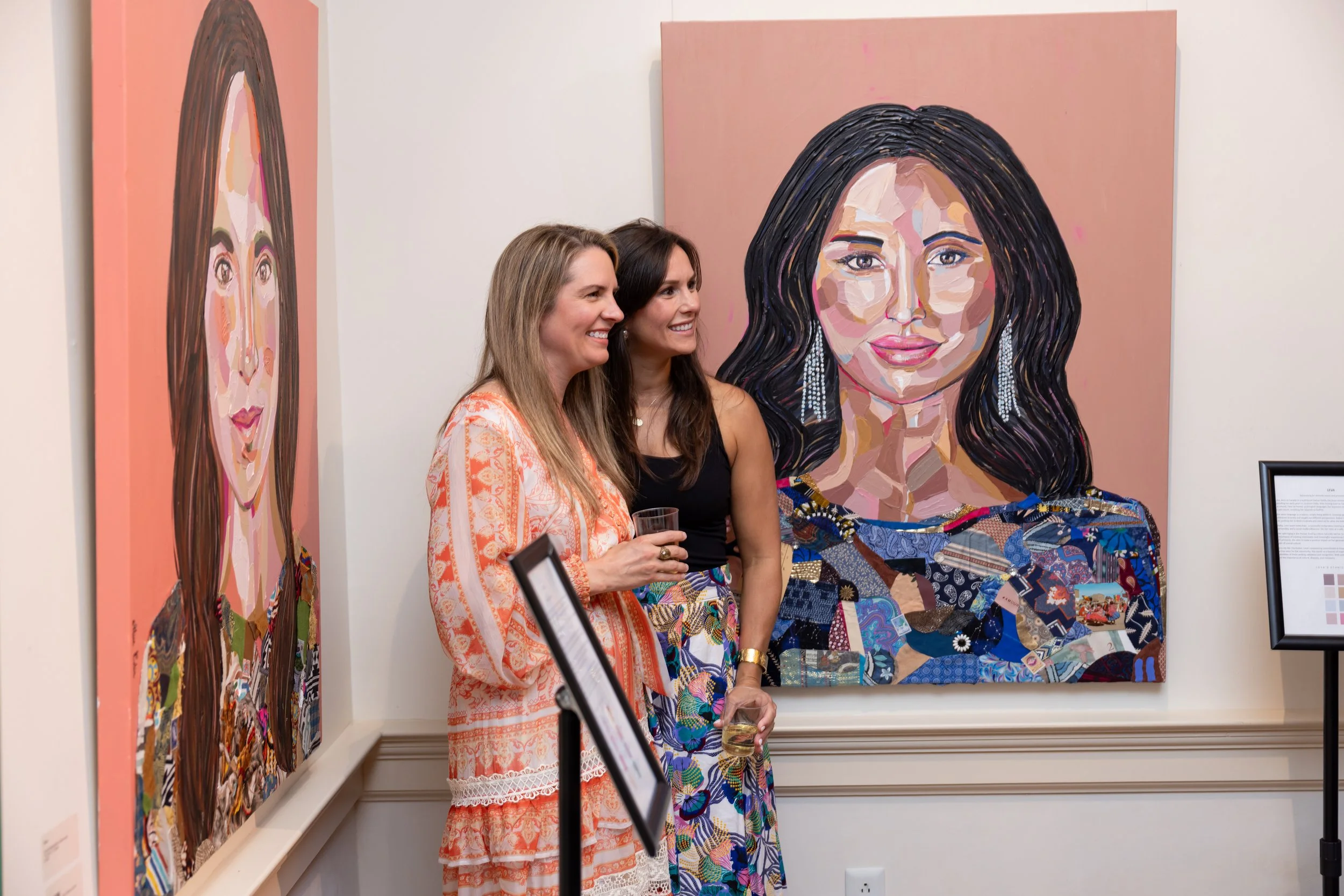 Two women standing together at an art gallery, observing colorful portrait paintings of women on the wall, with one woman holding a glass of wine.