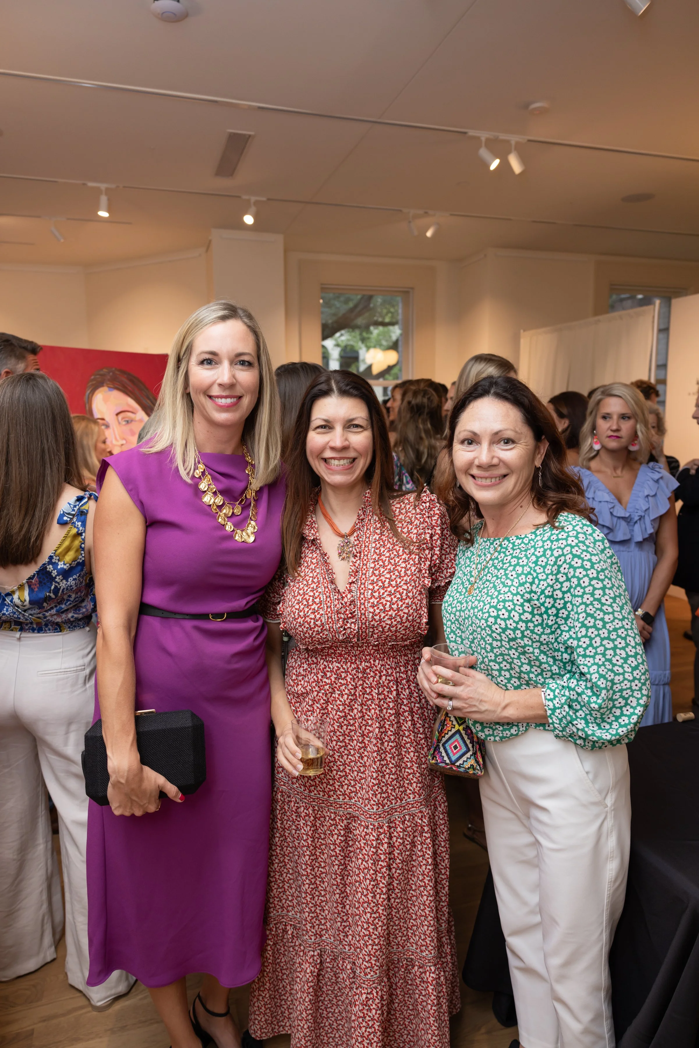 Three women smiling at an indoor social gathering. The woman on the left is wearing a purple dress with a gold necklace and holding a black clutch. The woman in the middle is wearing a red patterned dress, smiling, and holding a glass of drink. The w
