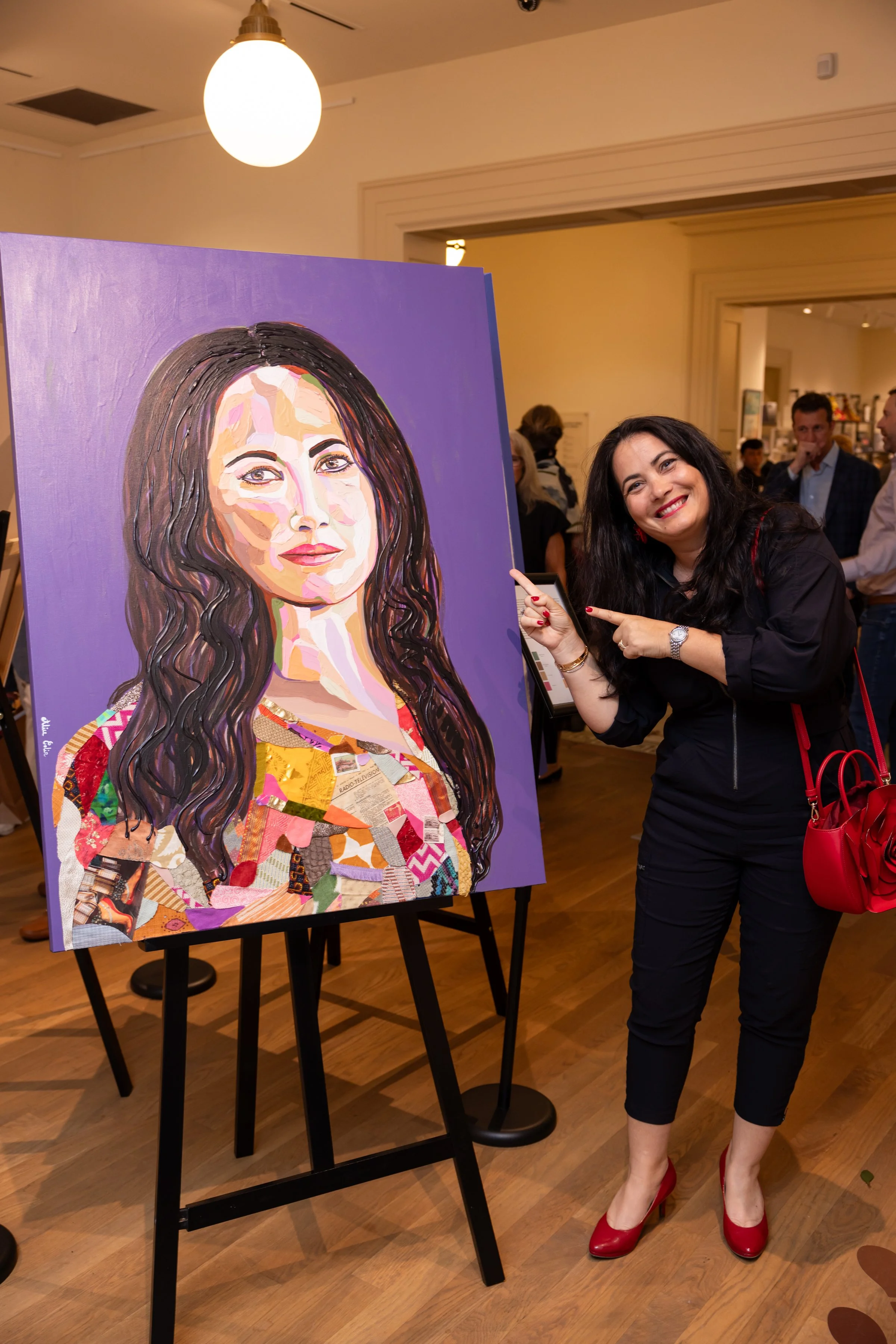 A woman with long black hair, red lipstick, wearing a black dress with red accents and red high heels, is pointing and smiling at a large colorful portrait painting of a woman with long dark hair, wearing a multicolored outfit, displayed on an easel 