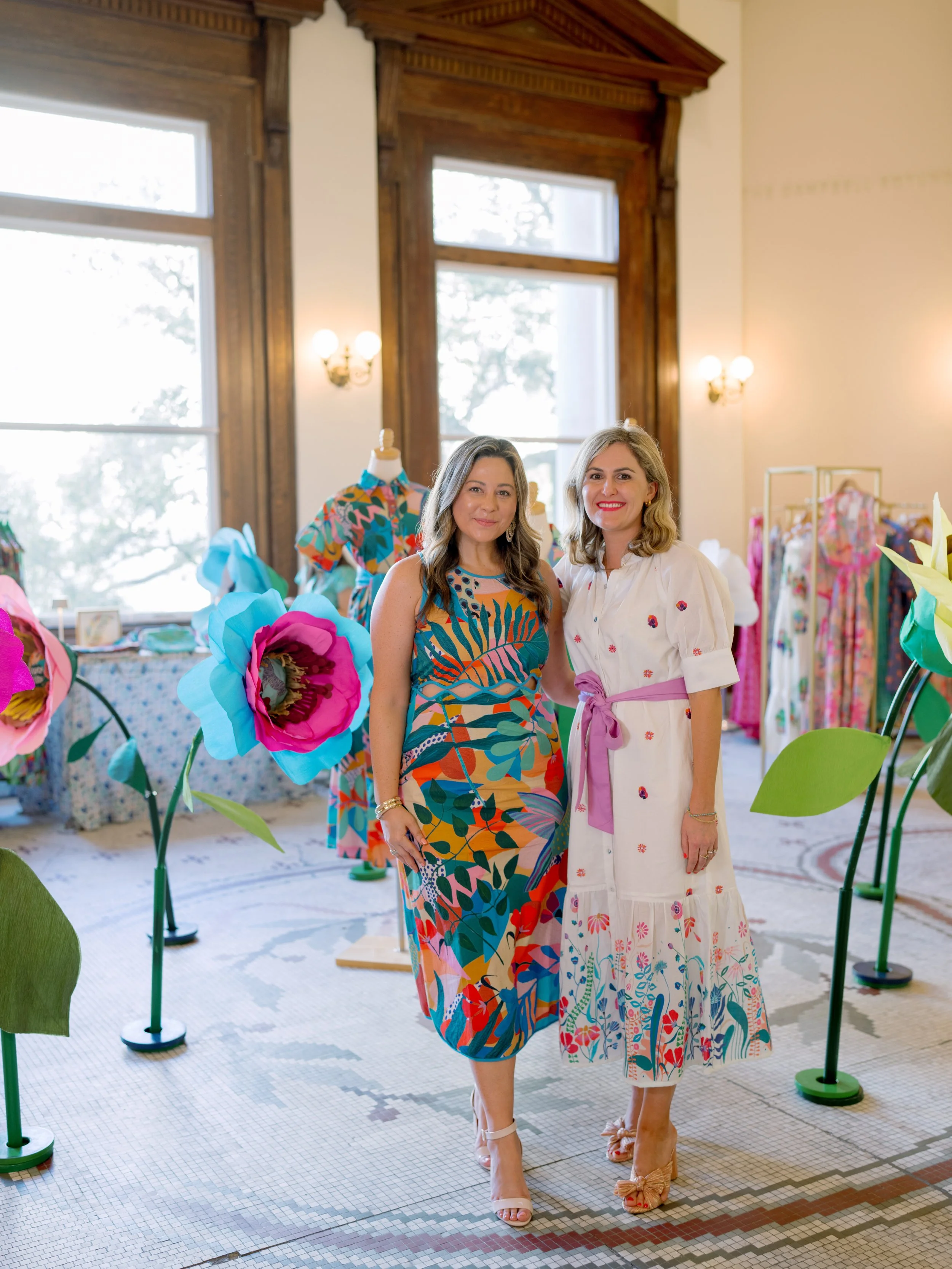 Two women standing inside a decorated room with large windows, wooden trim, and vintage lighting, surrounded by colorful large paper flower decorations and mannequins dressed in patterned clothing.