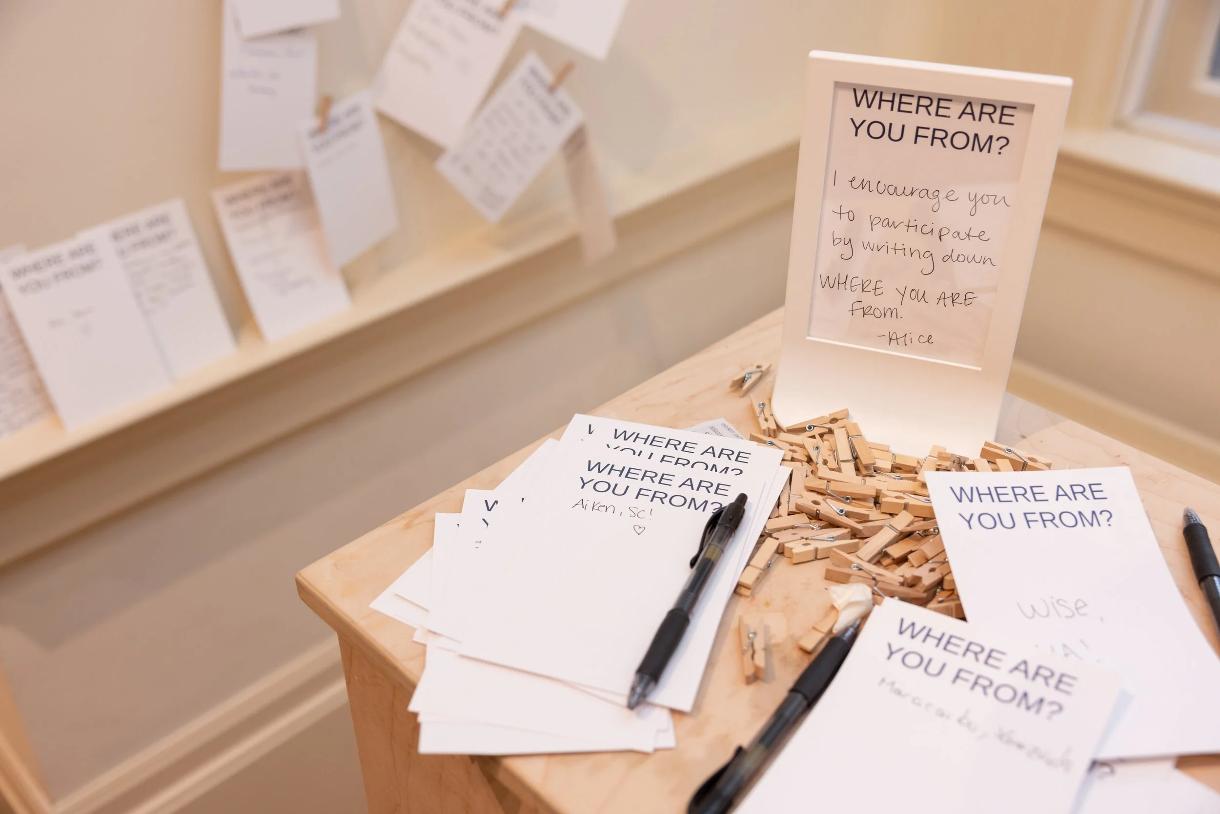 A table with printed sheets, black markers, and wooden clothespins. A sign on the table reads "WHERE ARE YOU FROM? I encourage you to participate by writing down WHERE YOU ARE from." In the background, there are more signs and papers attached to a wa