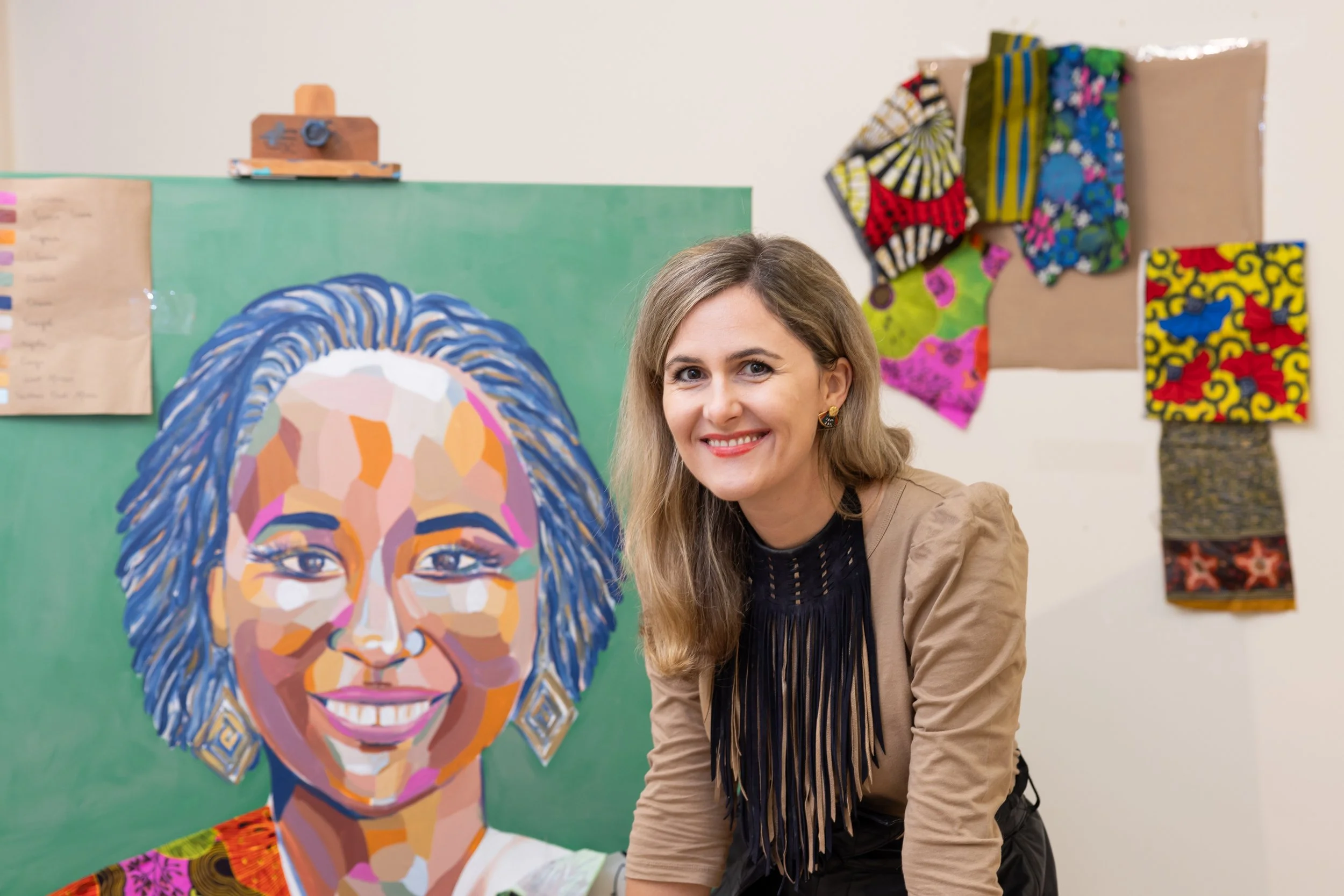 A woman with long blonde hair and earrings smiling at the camera indoors, with colorful artwork and fabric pieces hanging on the wall behind her.