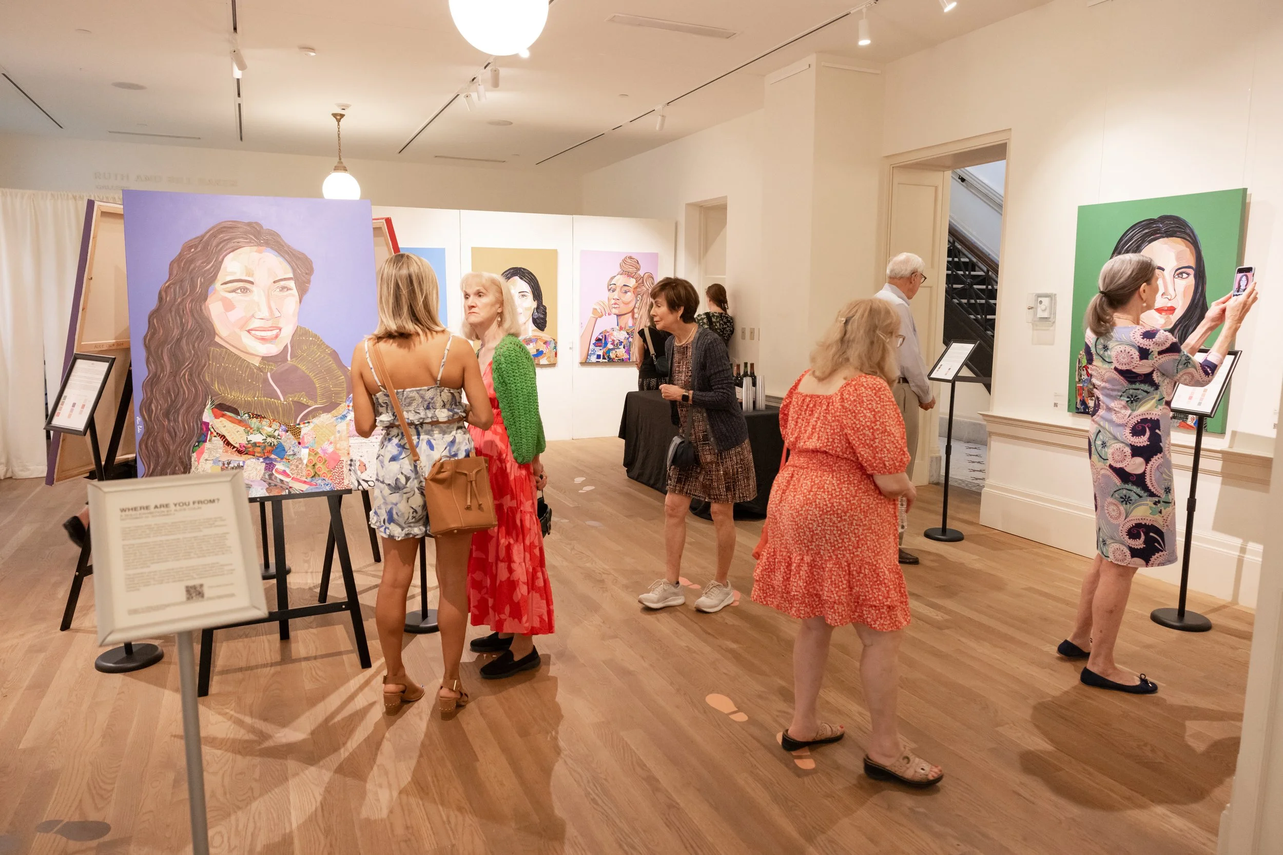 An art gallery with colorful portrait paintings of women displayed on white walls. Several visitors are observing the artworks, some are taking photos, and others are engaged in conversation. The room has wooden floors and soft lighting.