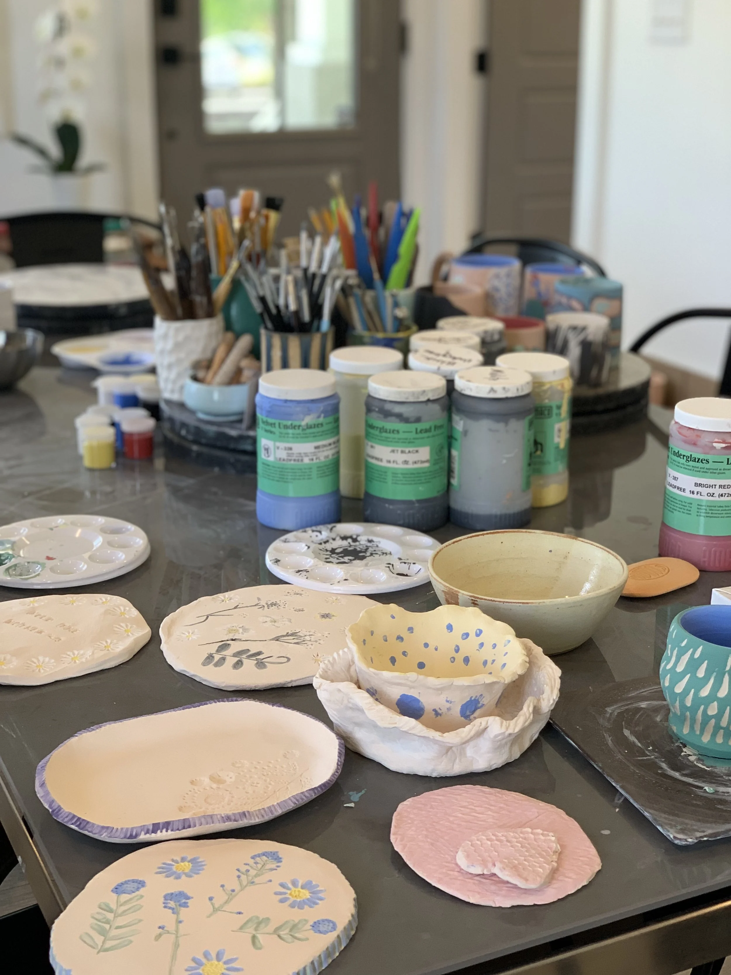 A workspace with various painted ceramic plates, bowls, and other pottery items on a table. There are jars of paint, paint brushes, and art supplies in the background.