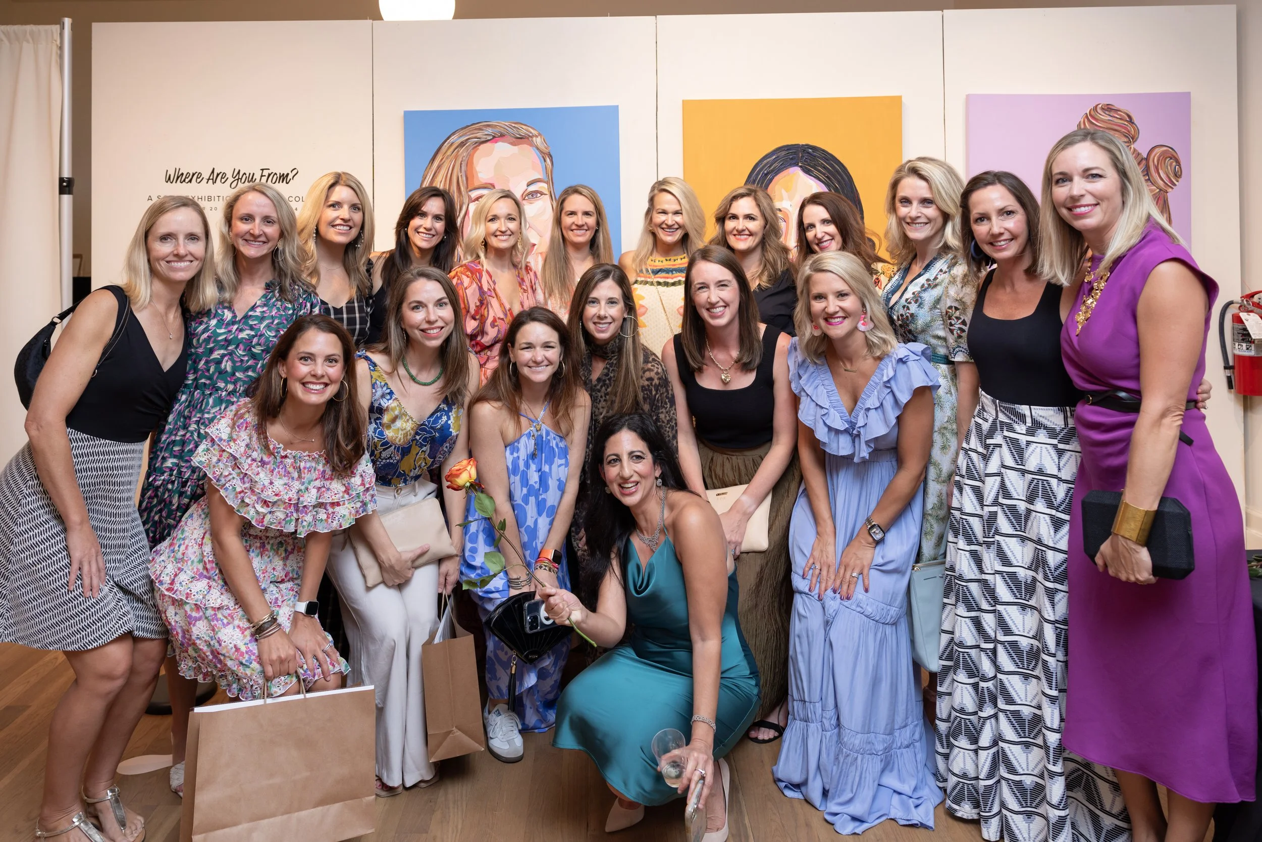 A group of women posing together at an art exhibition, standing in front of colorful portrait paintings.