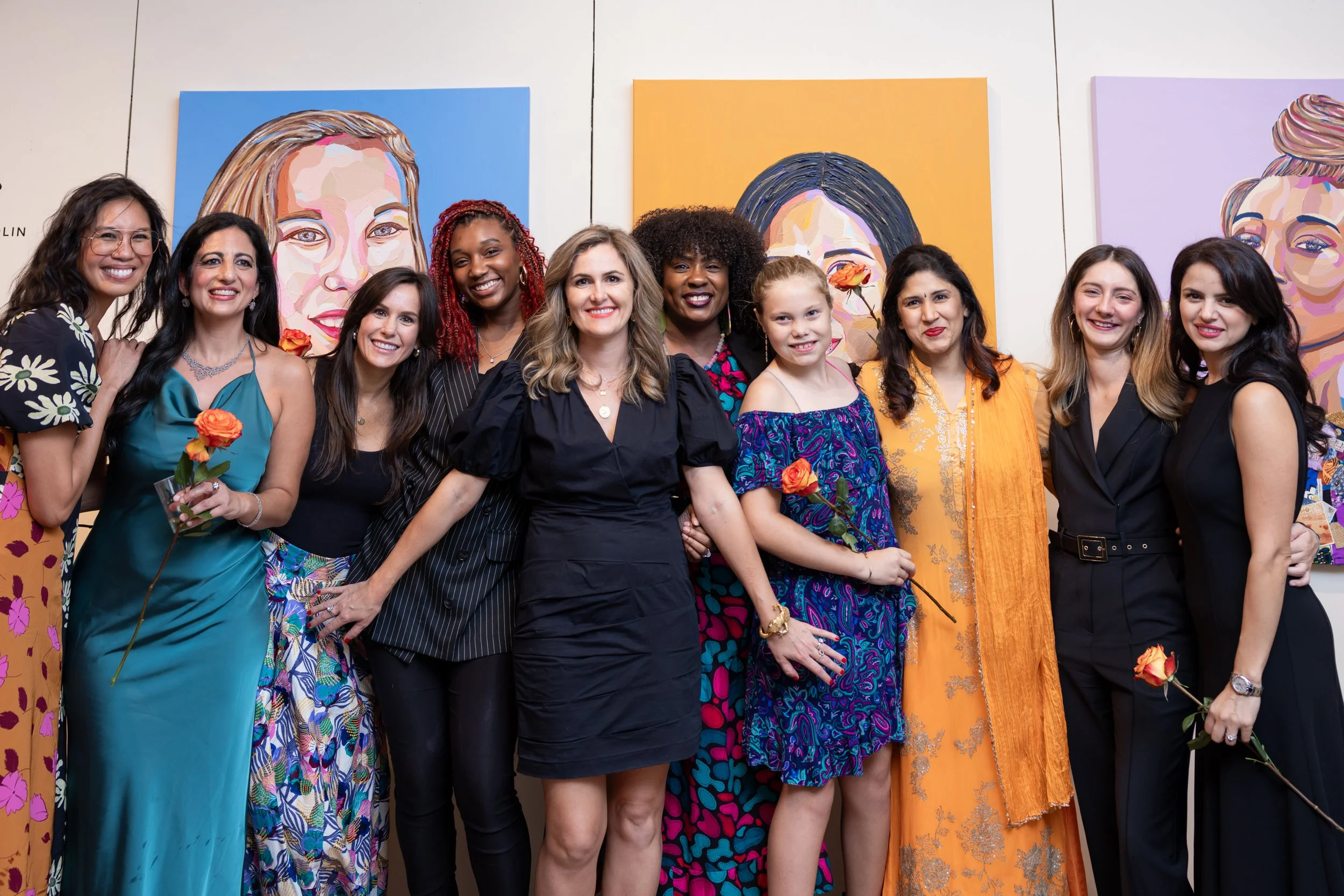 Group of diverse women at an art gallery, standing in front of colorful portrait paintings, smiling, some holding flowers.