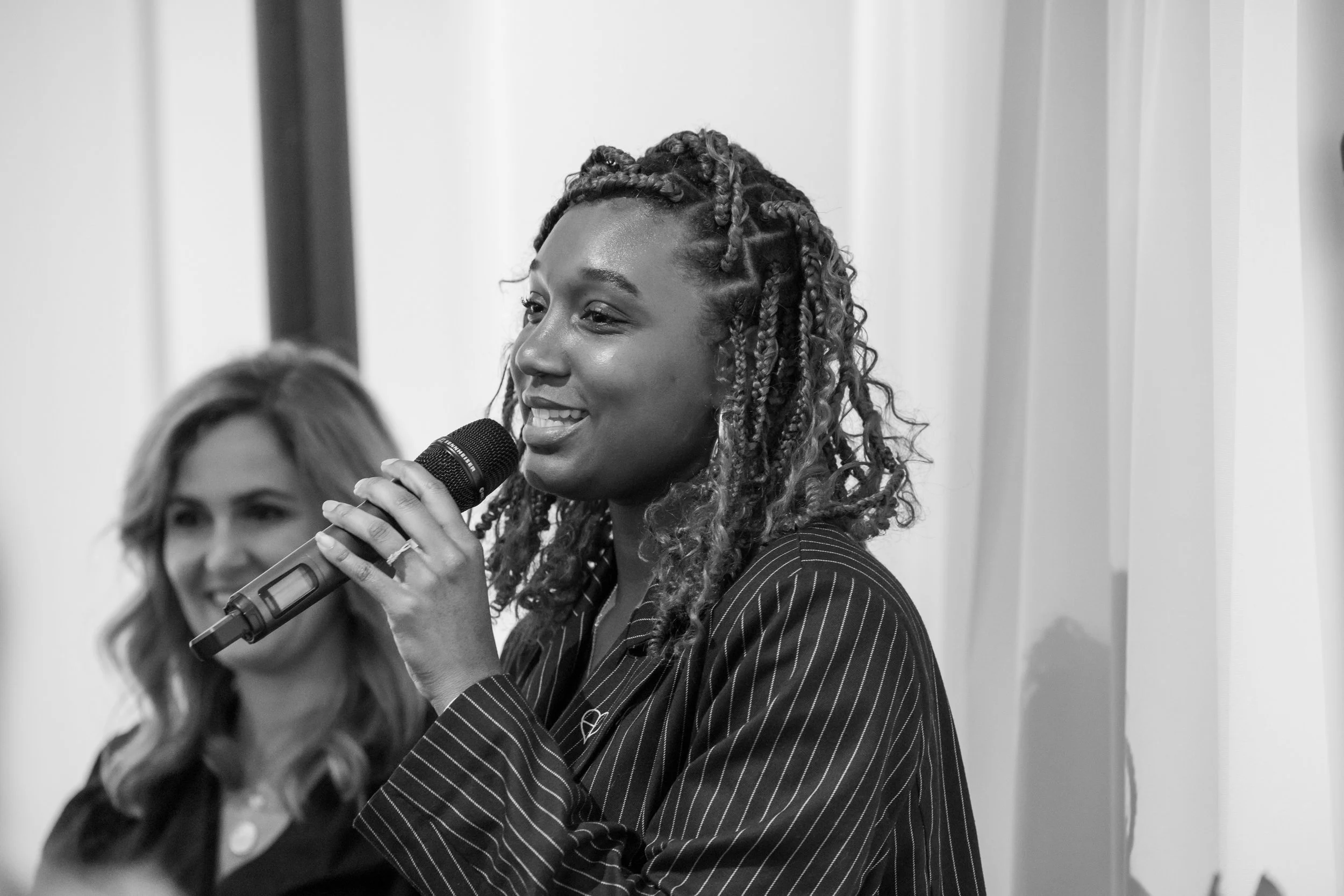 A Black woman with styled hair, wearing a pinstripe blazer, is smiling and speaking into a microphone. A woman with light hair and glasses is blurred in the background.