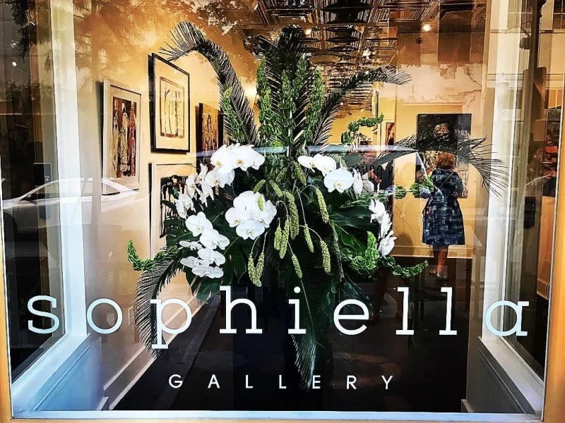 A window display with a large floral arrangement featuring white orchids and green foliage, with the words 'sophiela GALLERY' written at the bottom.