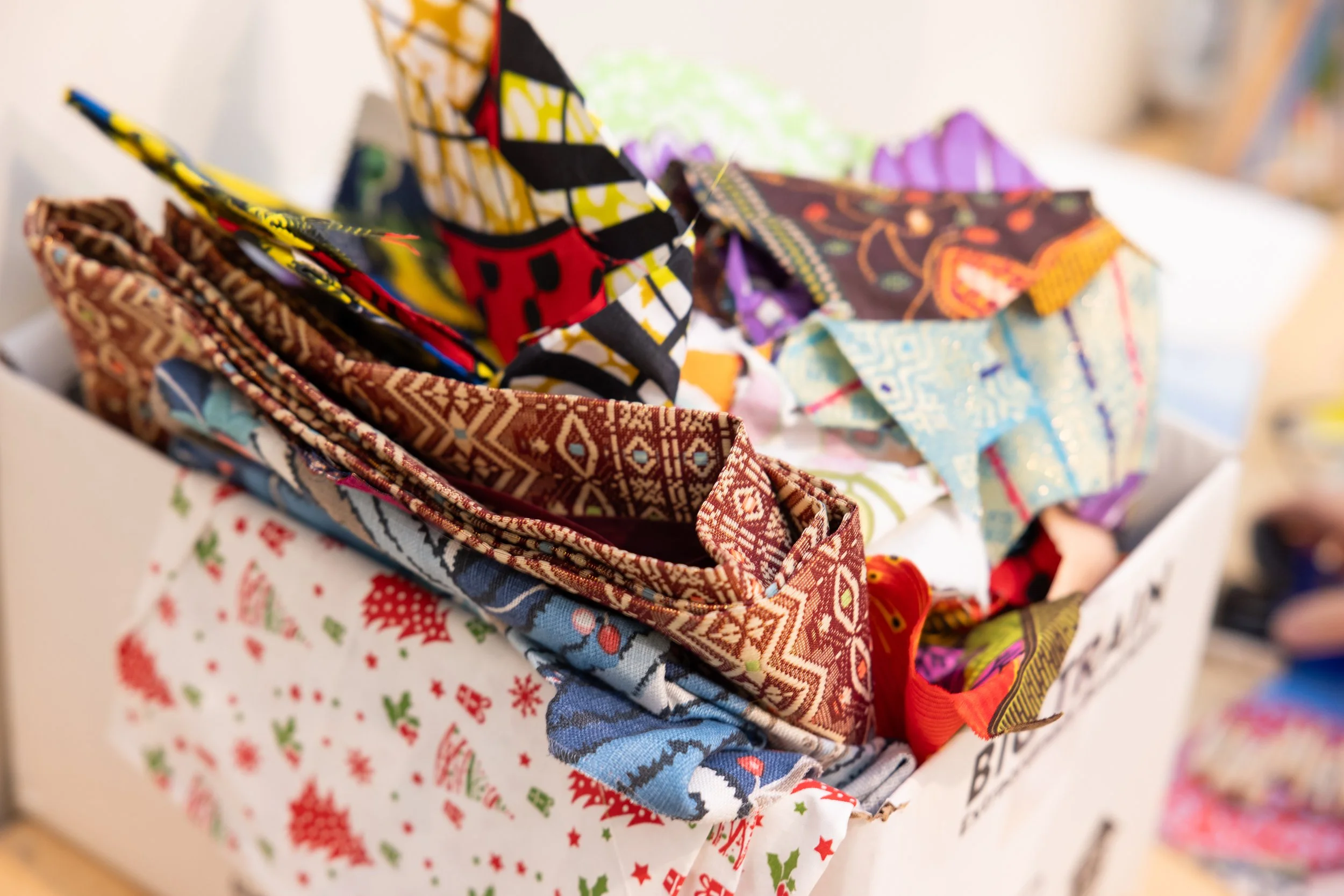 Close-up of various colorful fabric pieces with patterns, stacked and partially folded on a surface with holiday-themed wrapping paper underneath.