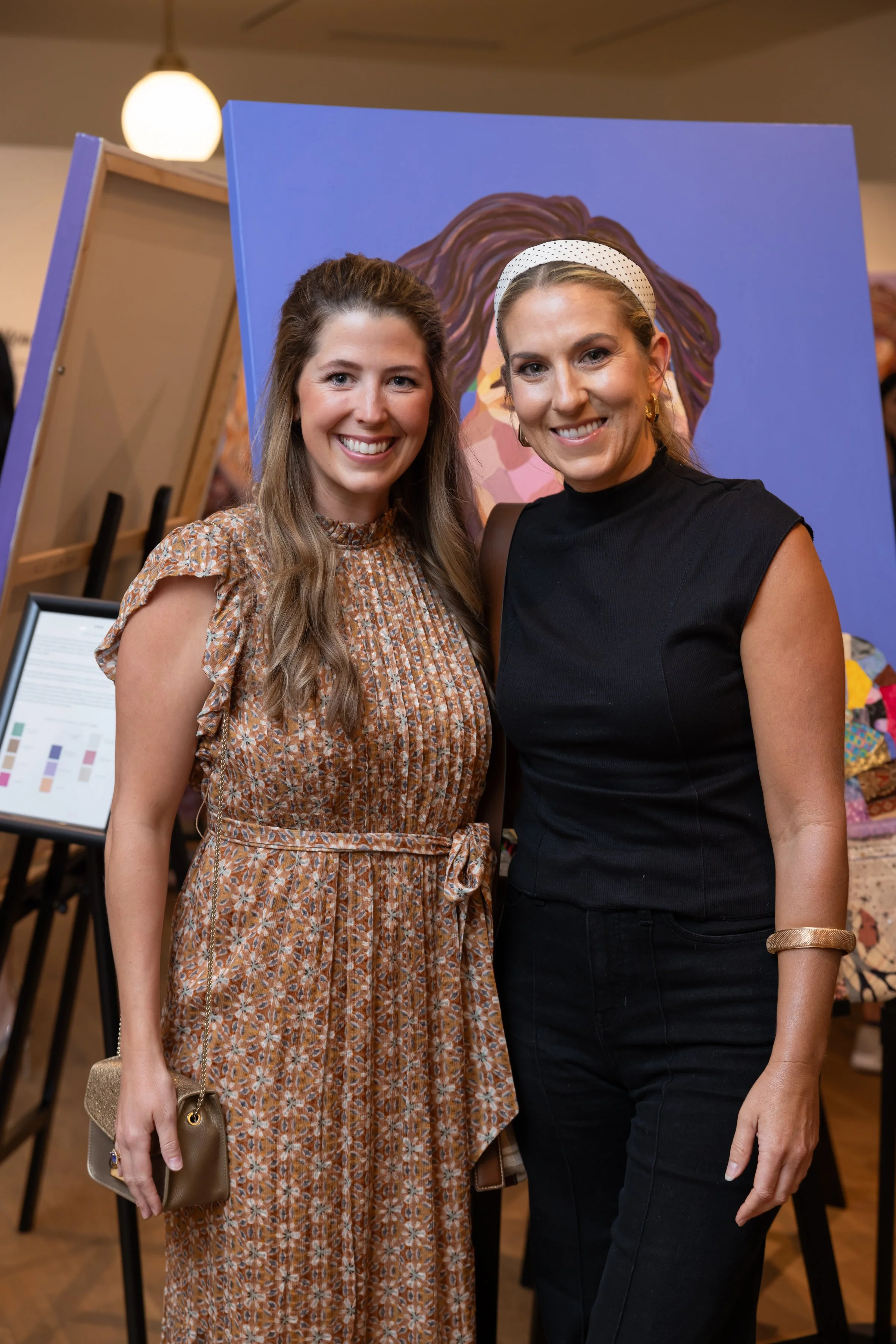 Two women standing close together, smiling at the camera, in front of a colorful portrait and display boards at an art event or gallery.