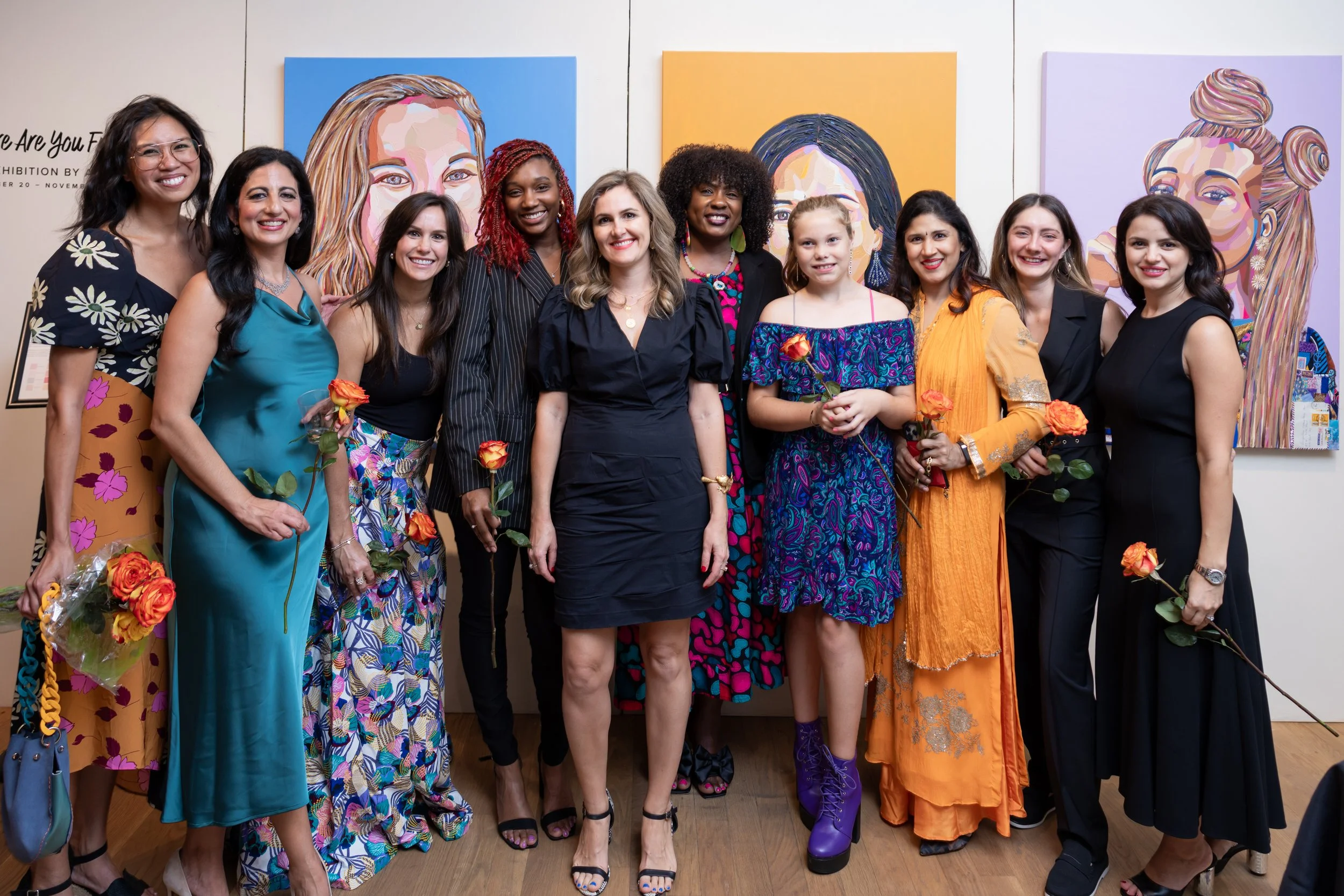 Group of women standing together at an art exhibit, holding roses, with colorful portrait paintings in the background.