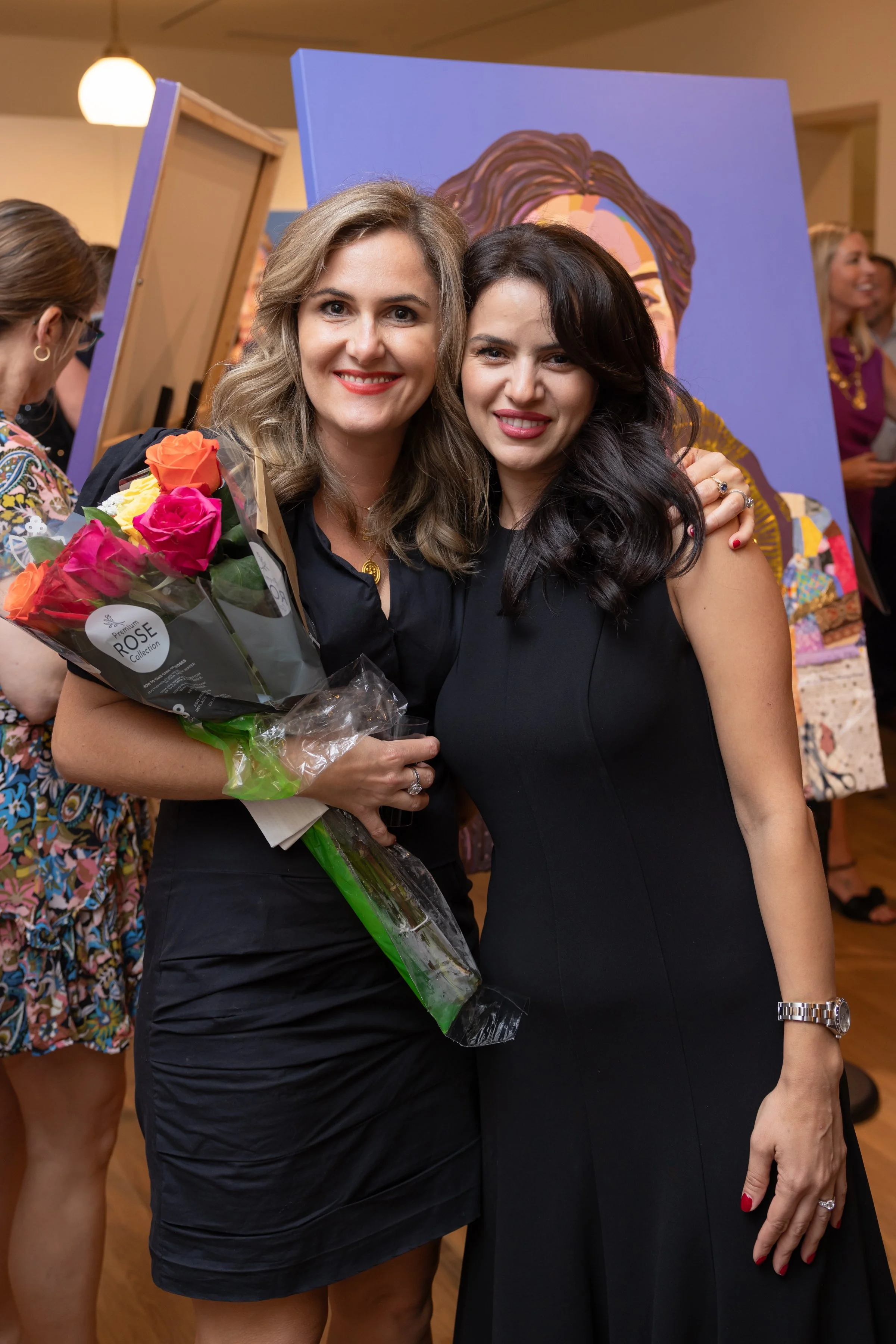 Two women standing close together smiling, one holding a bouquet of pink, orange, and yellow roses. They are dressed in black and are at an indoor event with people in the background and artwork displayed.