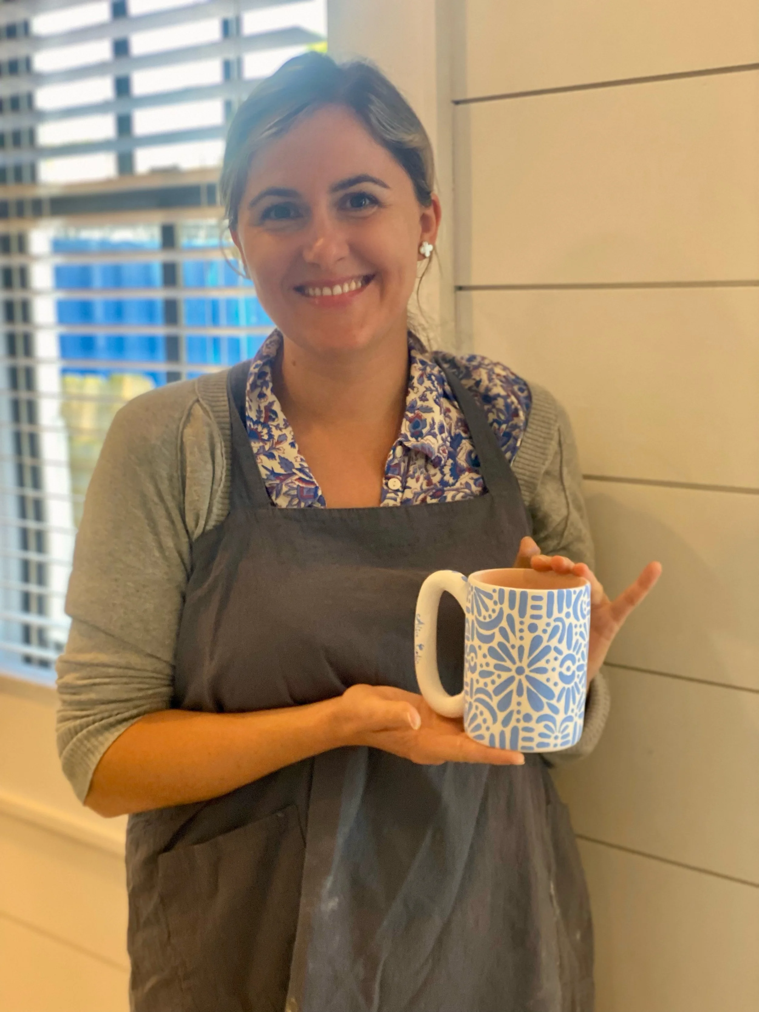 Smiling woman holding a white mug with blue floral pattern, standing indoors near a window with blinds.