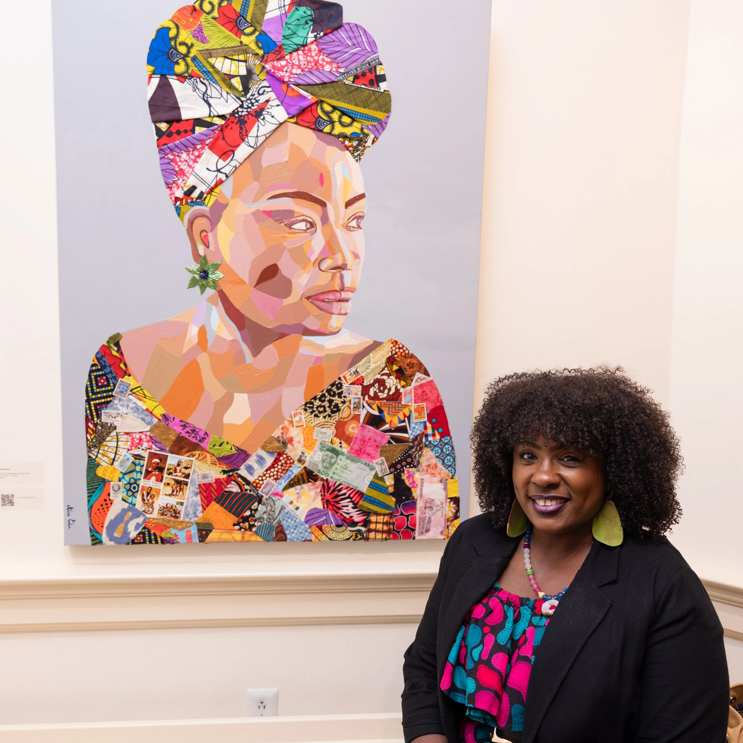 A woman with curly hair and earrings smiling in front of a colorful artwork depicting a woman wearing a head wrap and earrings, dressed in vibrant, patterned clothing.
