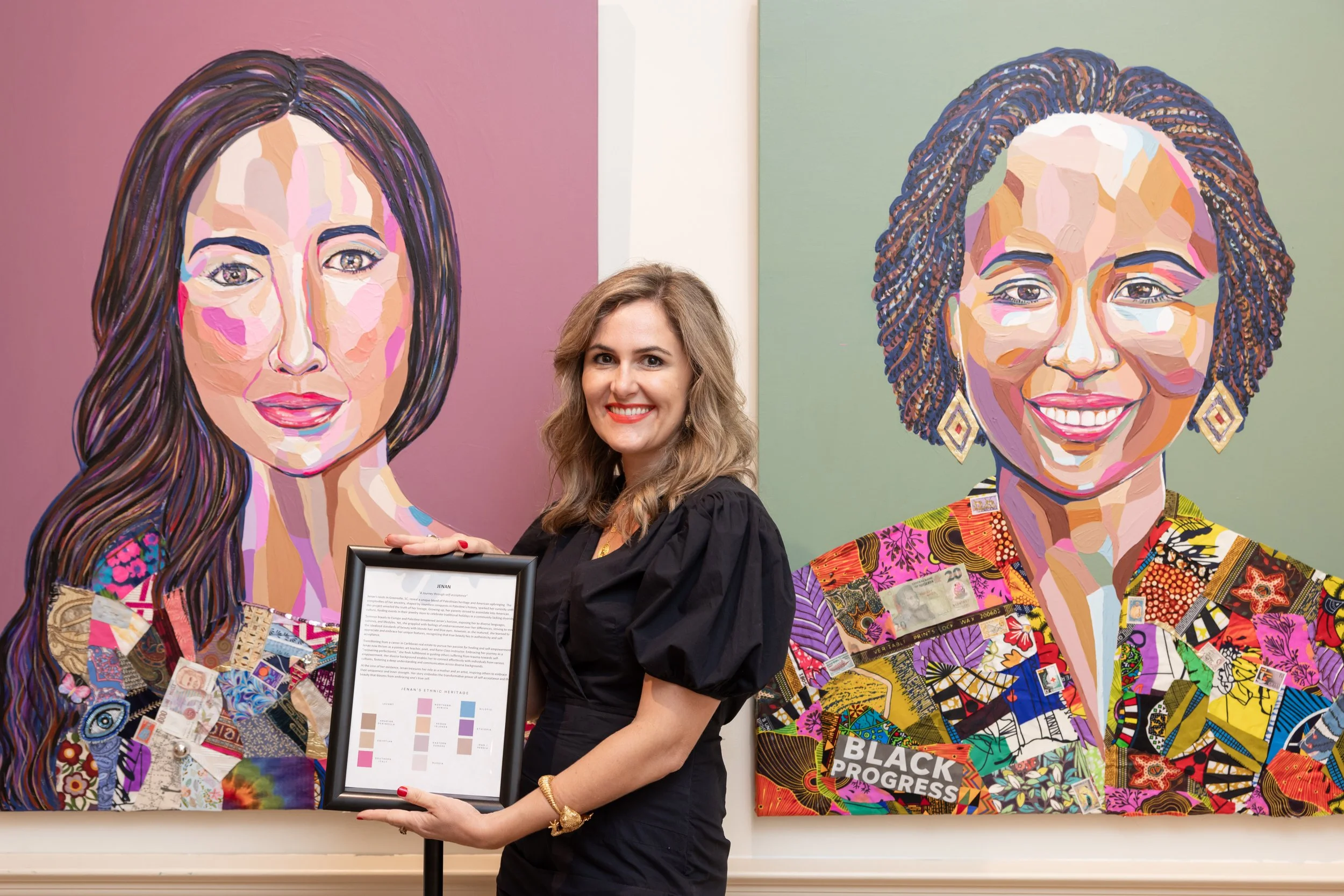 A woman with wavy light brown hair, wearing a black top and red lipstick, standing in front of two colorful portraits of women on a wall. She holds a framed document and smiles at the camera.